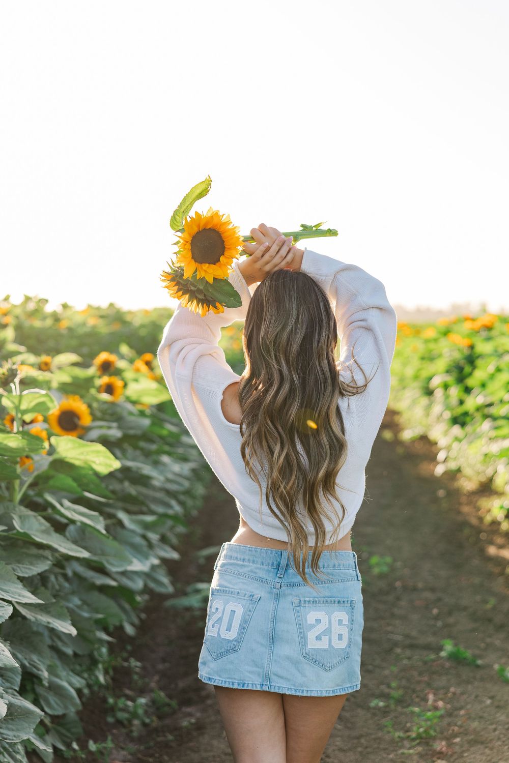 Woman in denim skirt holds sunflower overhead in golden sunflower field.