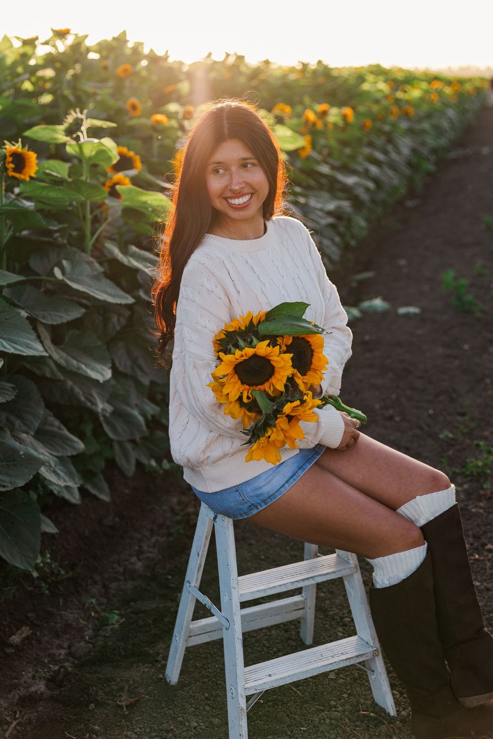 A person sits on a white chair in a sunflower field during golden hour wearing a cream sweater.