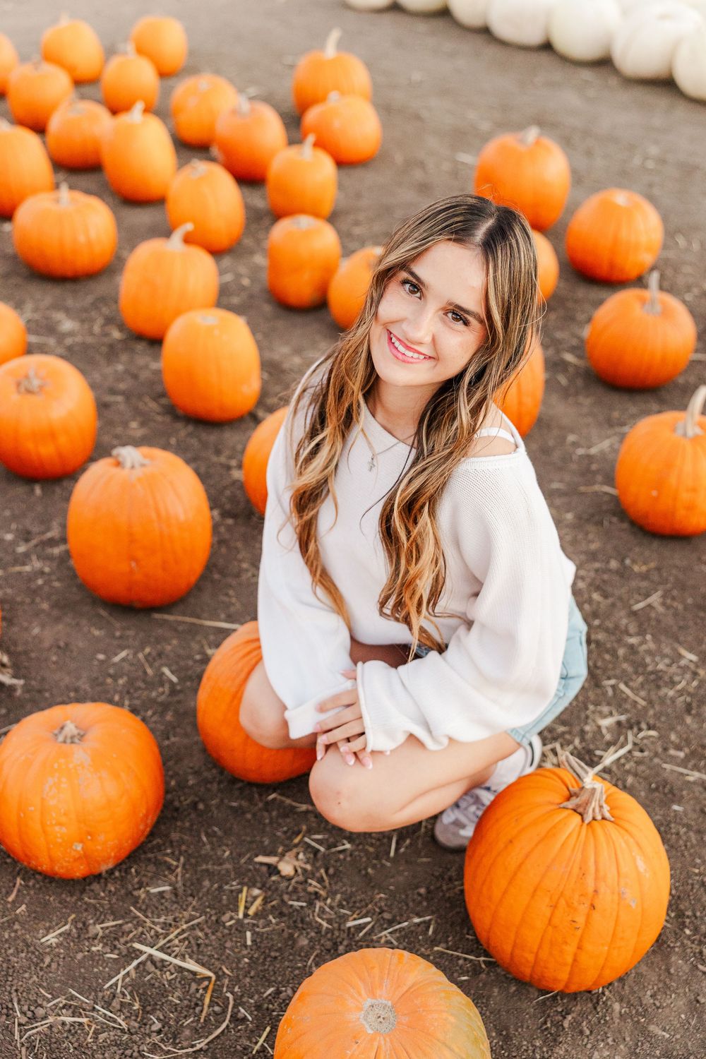 Woman in white sweater sitting among orange pumpkins at a fall harvest patch.