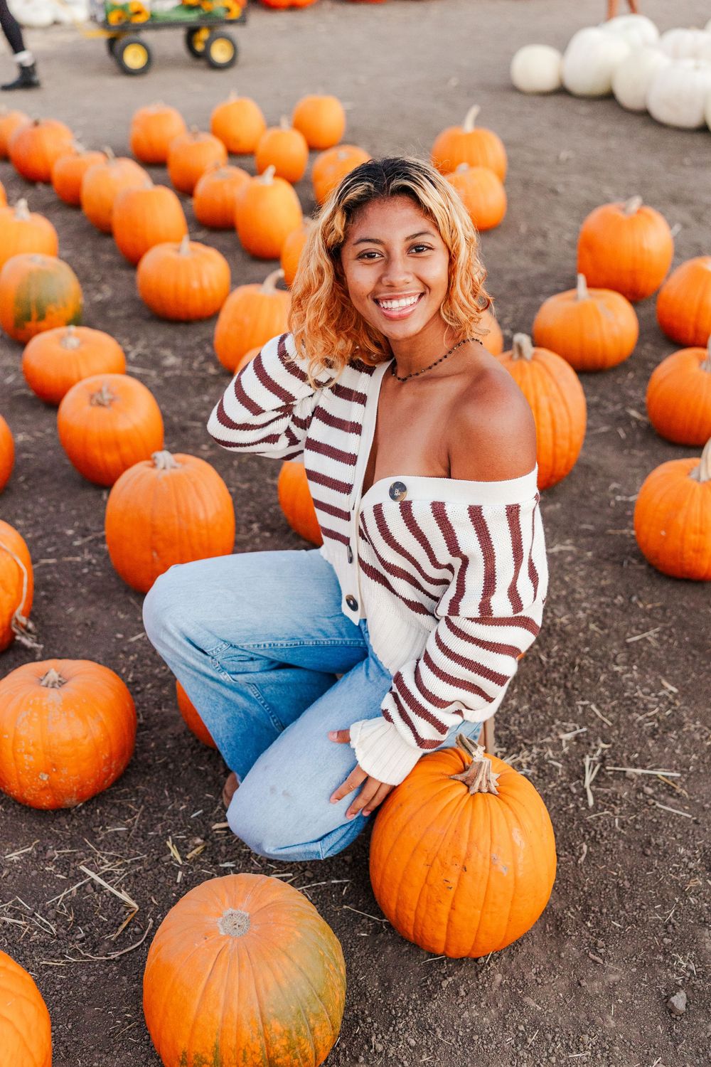 A person in a striped sweater sits among orange pumpkins at a fall pumpkin patch.