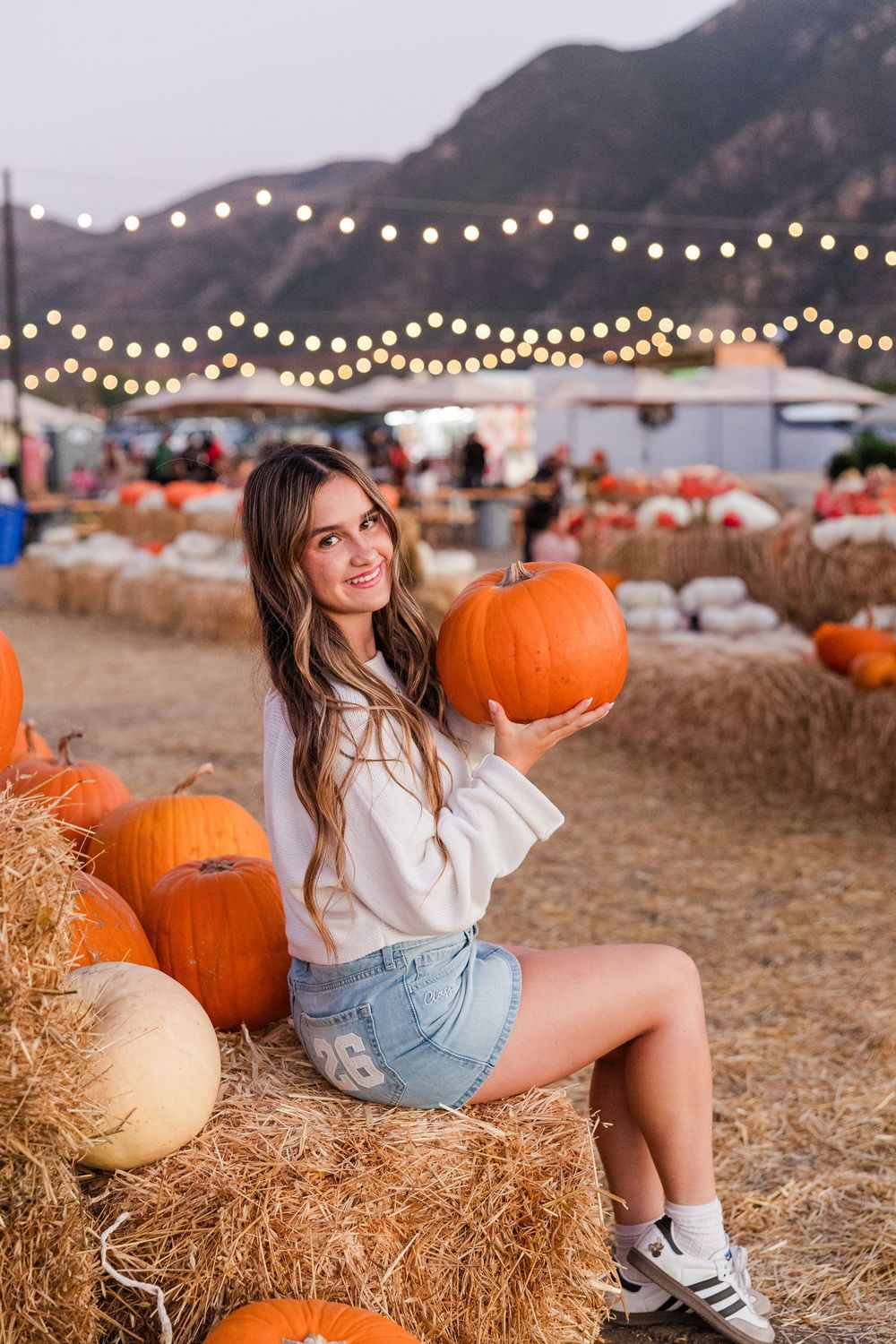 Rustic pumpkin patch setting with hay bales, string lights, and mountain backdrop at sunset.