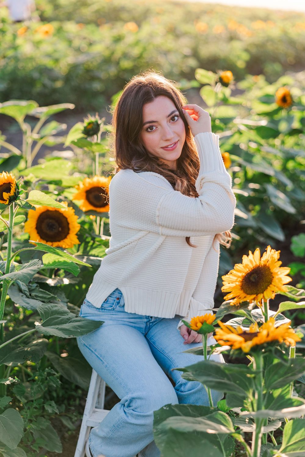 Woman in white sweater and jeans poses gracefully in a vibrant sunflower field during golden hour.