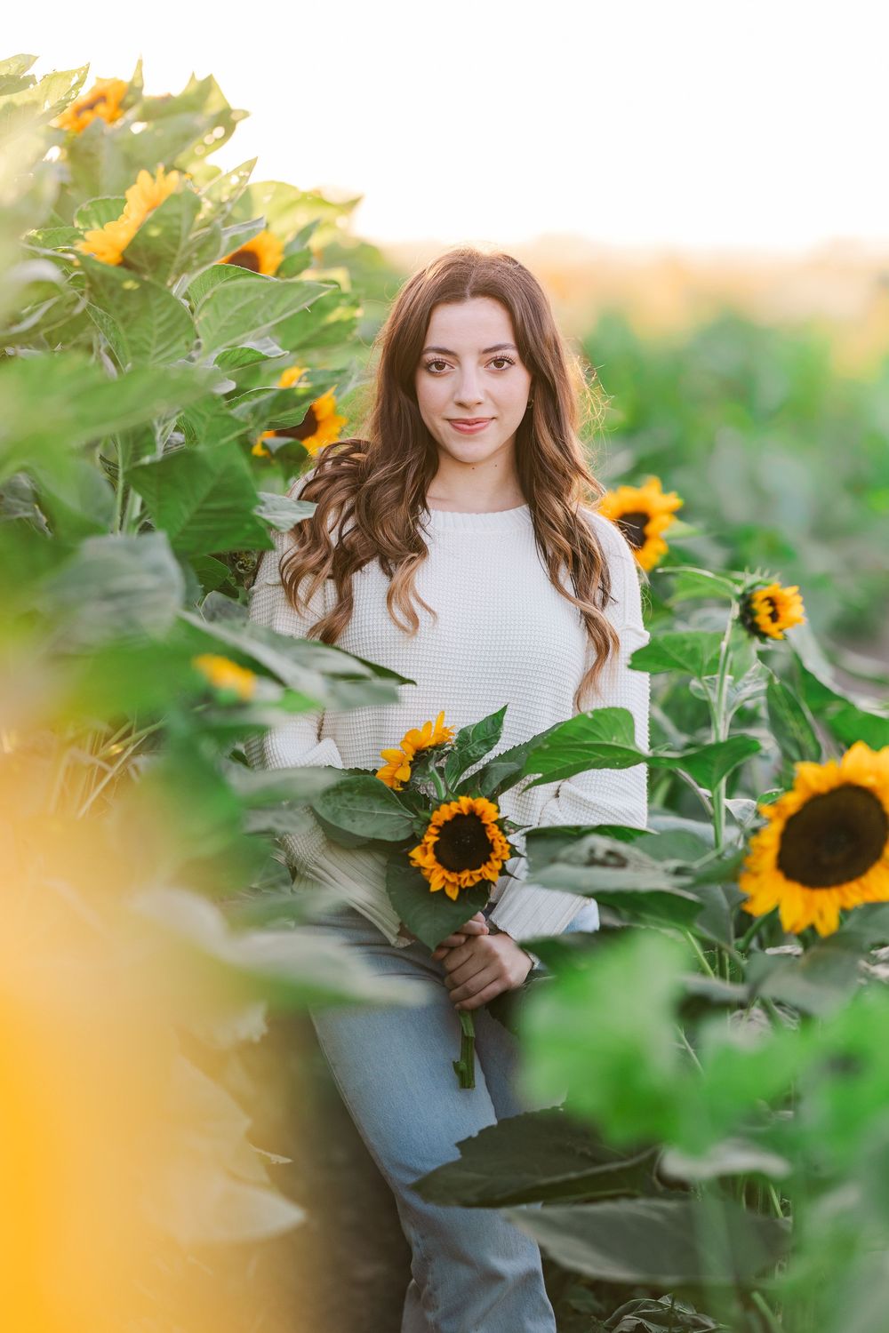 A person in a white sweater stands among blooming sunflowers in a garden during golden hour.
