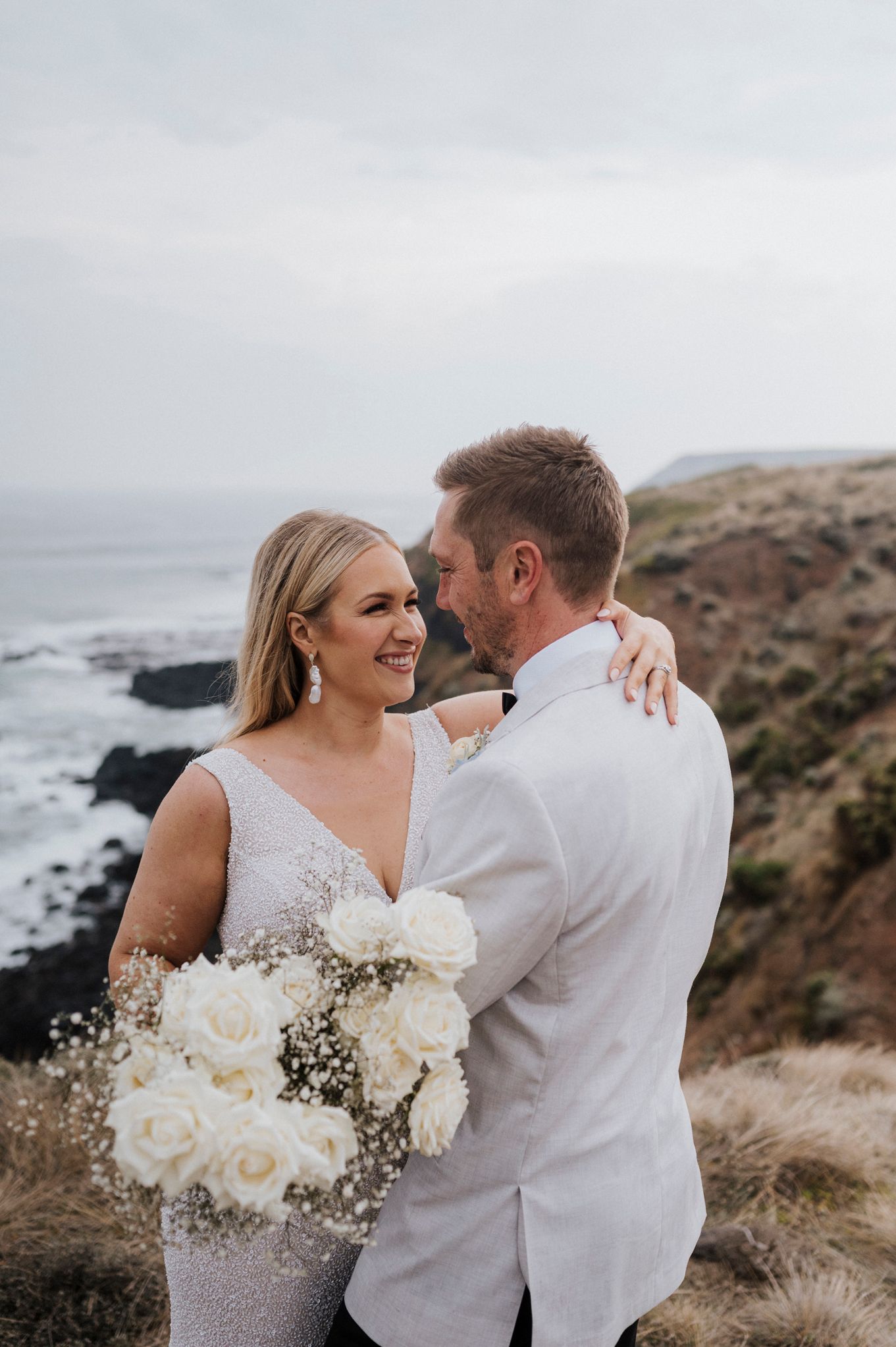 A couple embraces on a dramatic coastal cliff at sunset with crashing waves and rocky shoreline below.
