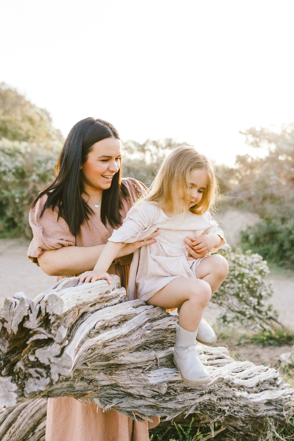 Two friends share joyful moments while sitting together on a weathered log in a sunlit outdoor setting.