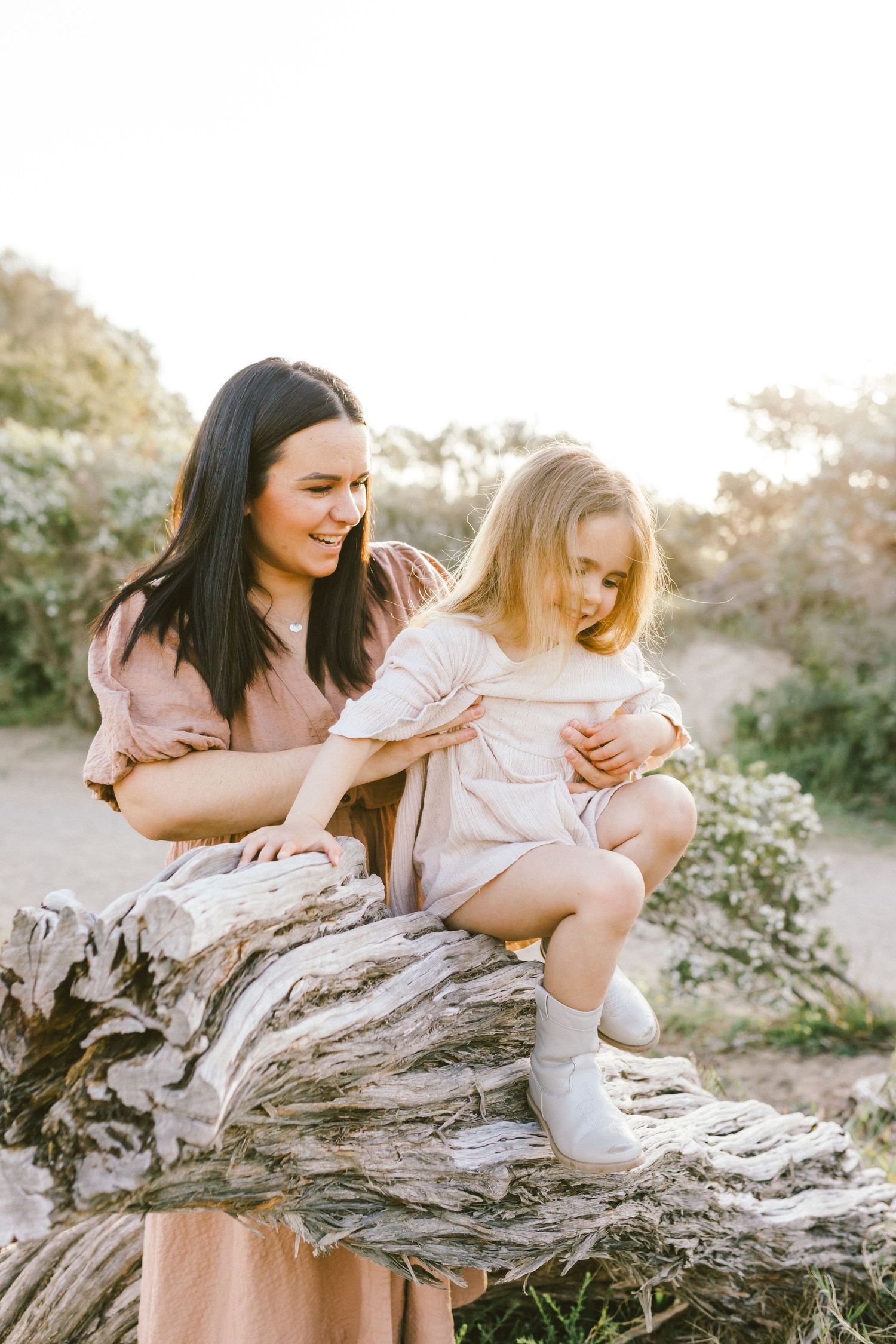 Two friends share joyful moments while sitting together on a weathered log in a sunlit outdoor setting.