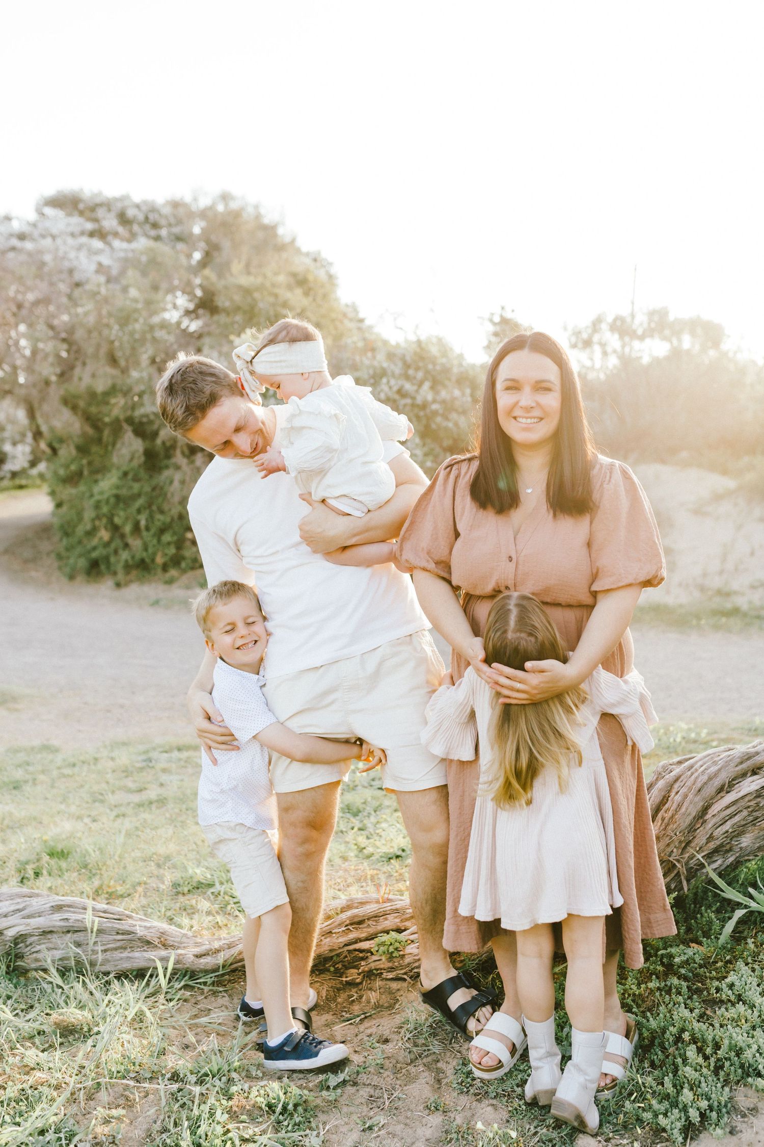 A family wearing light colored clothing poses together outdoors during golden hour in a natural setting.