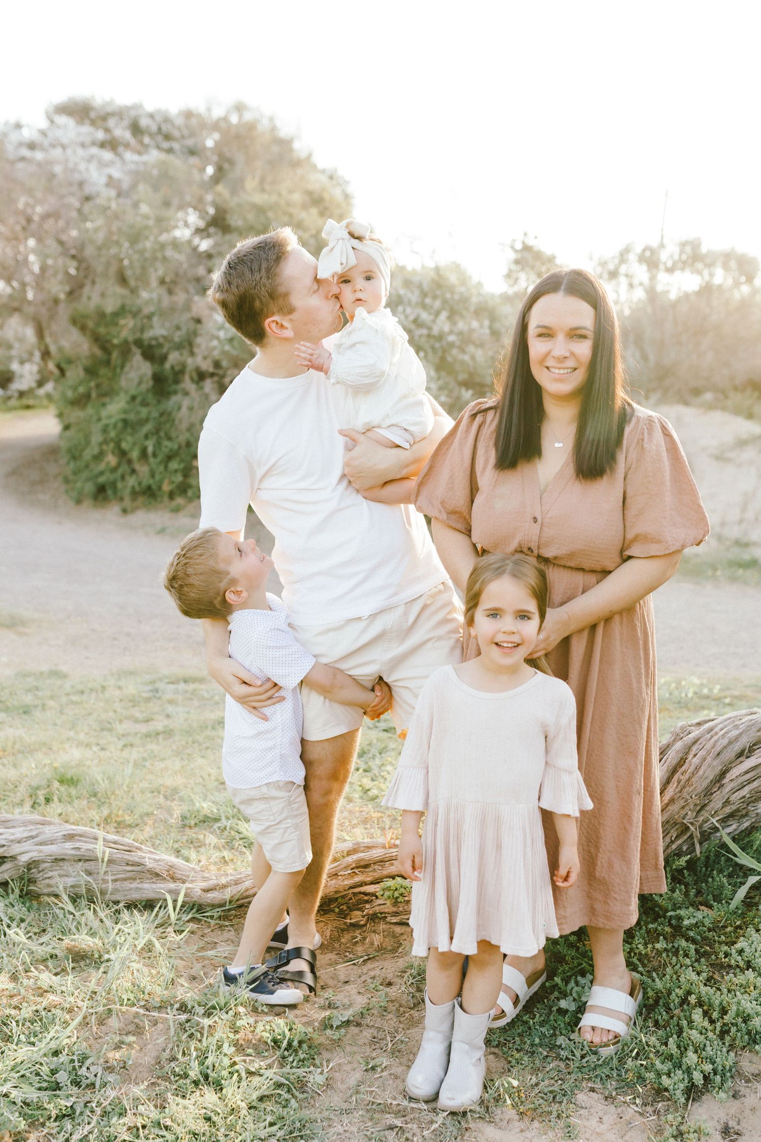 Family wearing neutral tones embraces and shares joyful moments together in a sunlit outdoor setting.