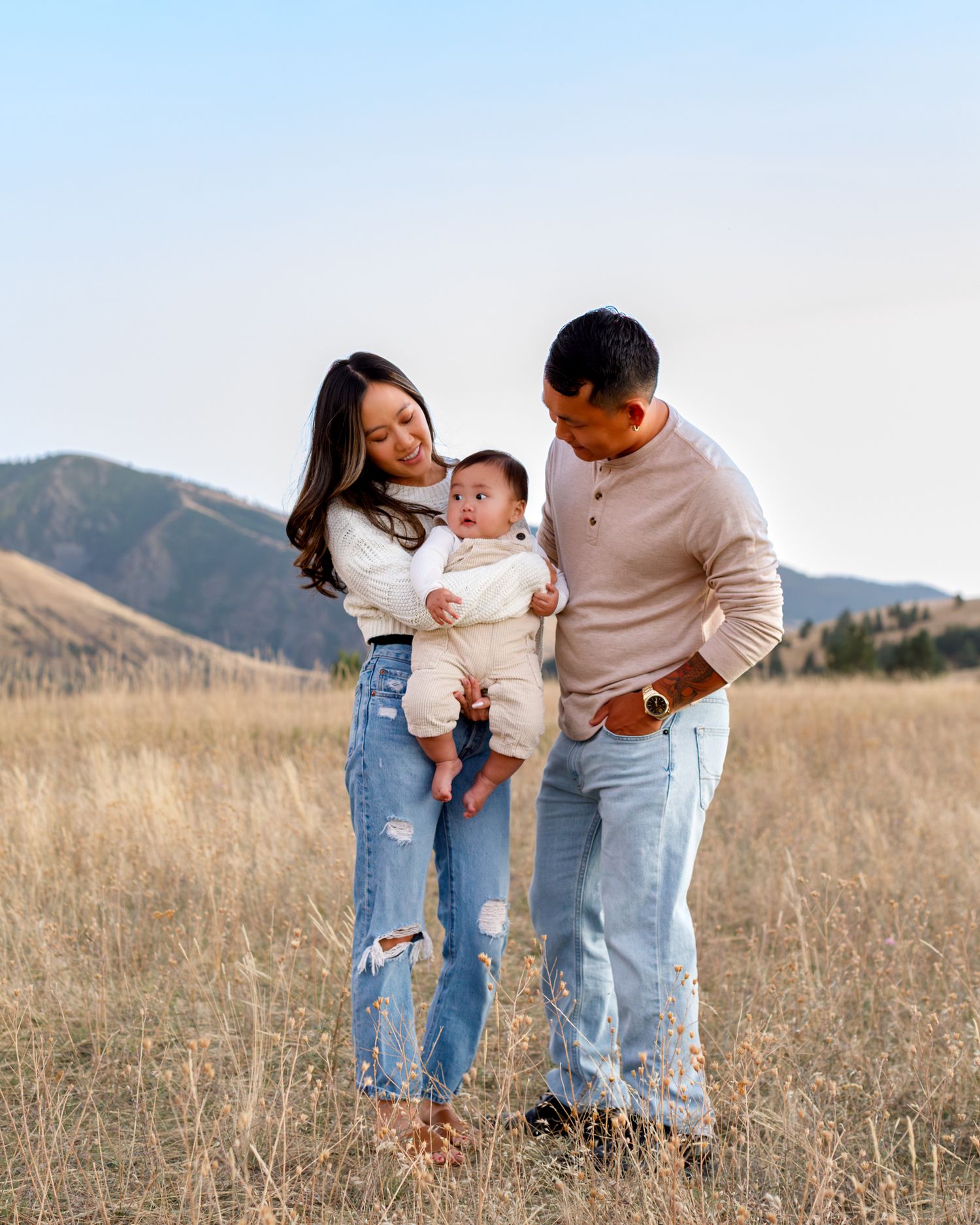 A couple gazes lovingly in a grassy meadow at sunset with mountains in the background with baby