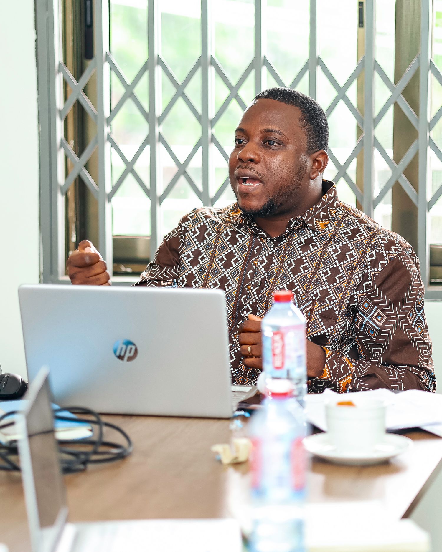 Professional in patterned shirt gesturing while speaking during business meeting with laptop.