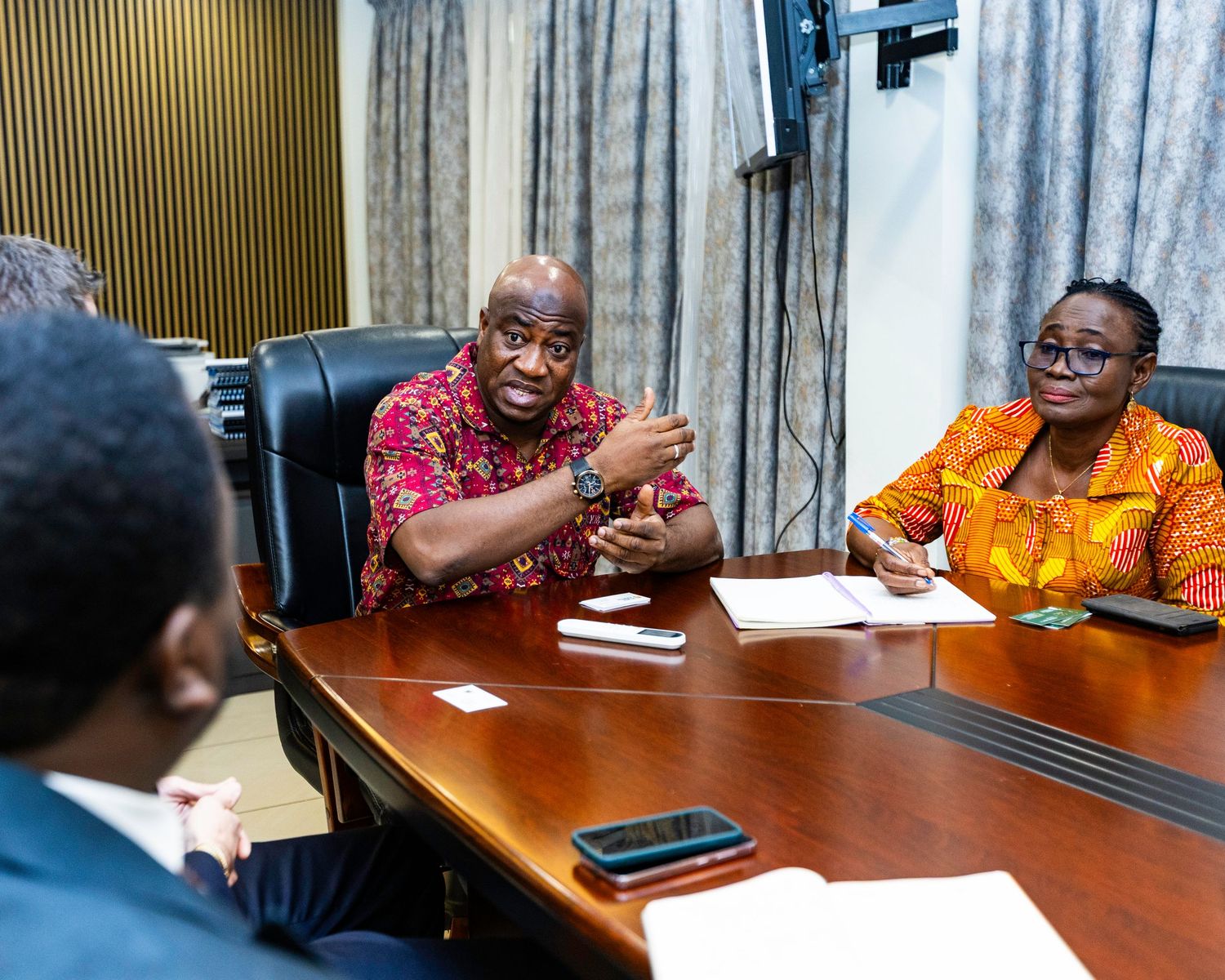 Business meeting participants engaged in discussion at conference table with wood paneling and curtains in background.