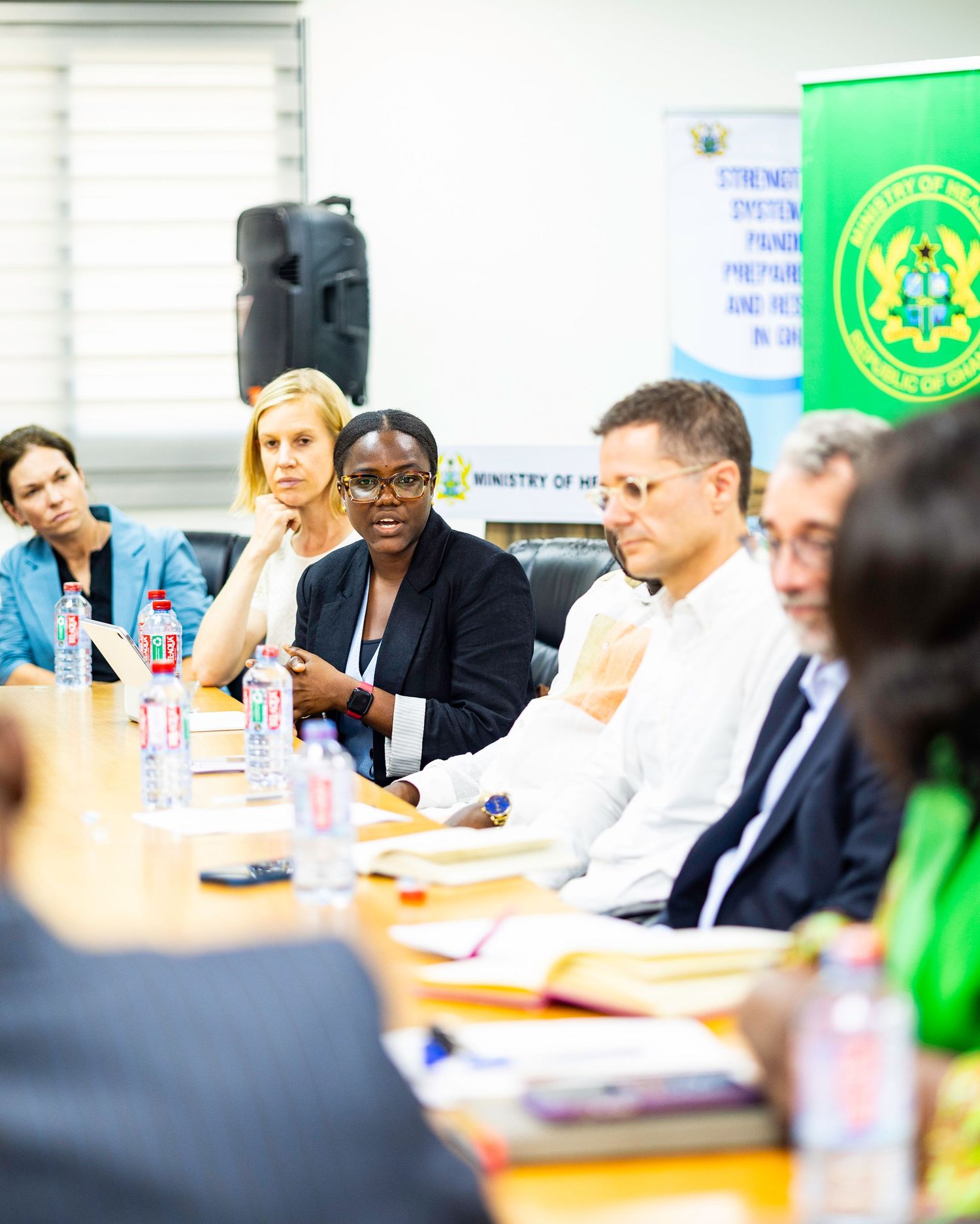 Conference room meeting with attendees seated around table with Ghana flag banner visible.