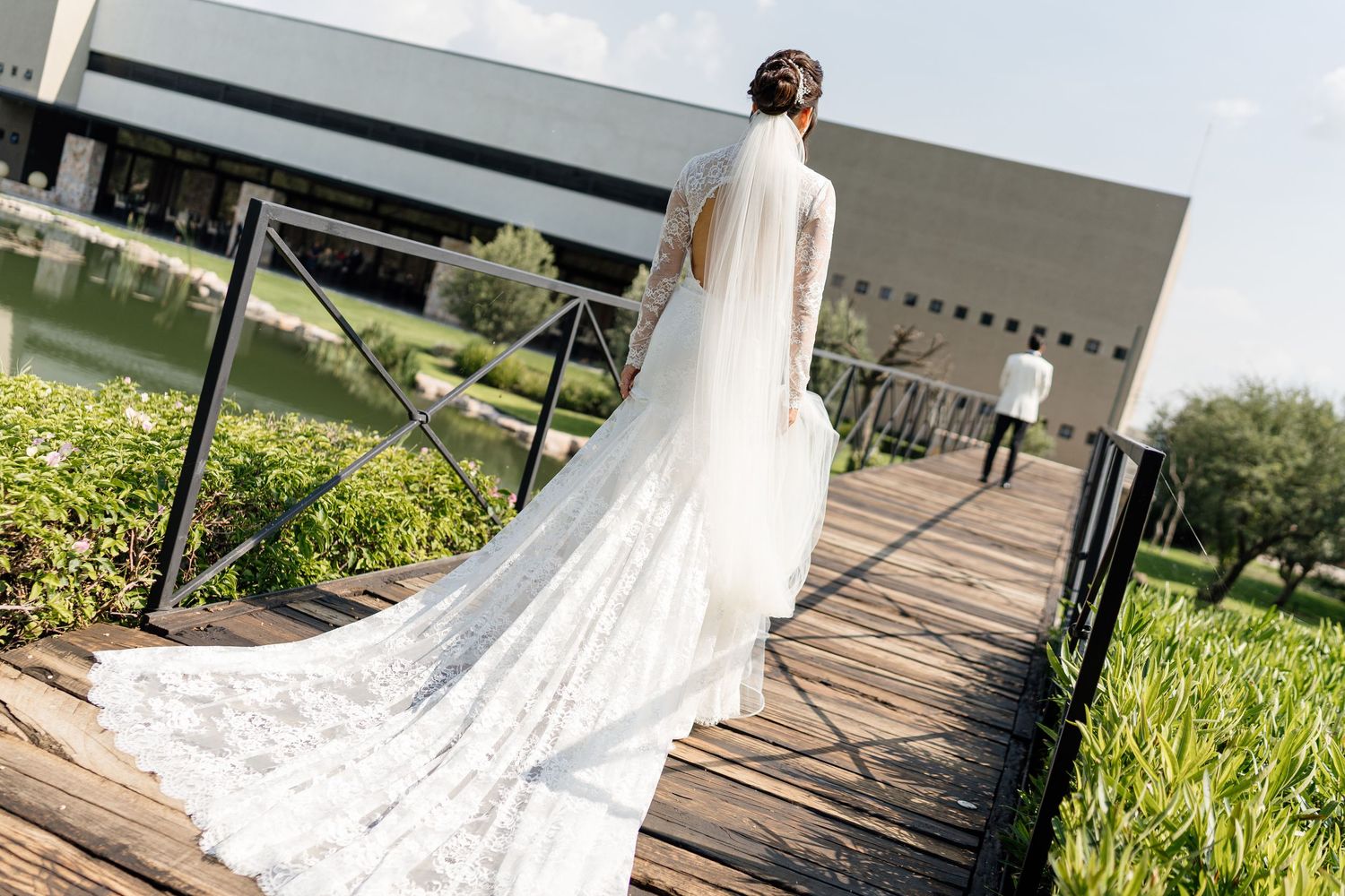 A bride in a flowing white lace wedding gown with cathedral train poses in a garden setting during a sunny day.