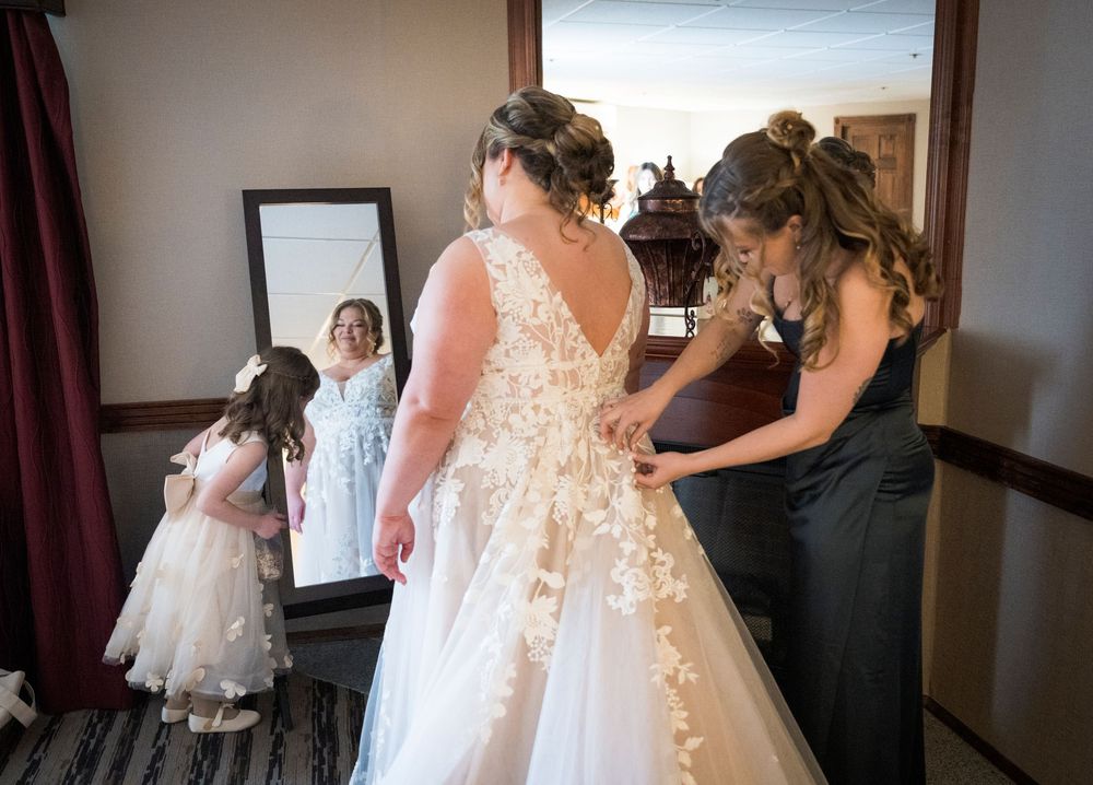 A wedding party helps adjust a beautiful lace wedding dress with floral appliques in a getting ready room.