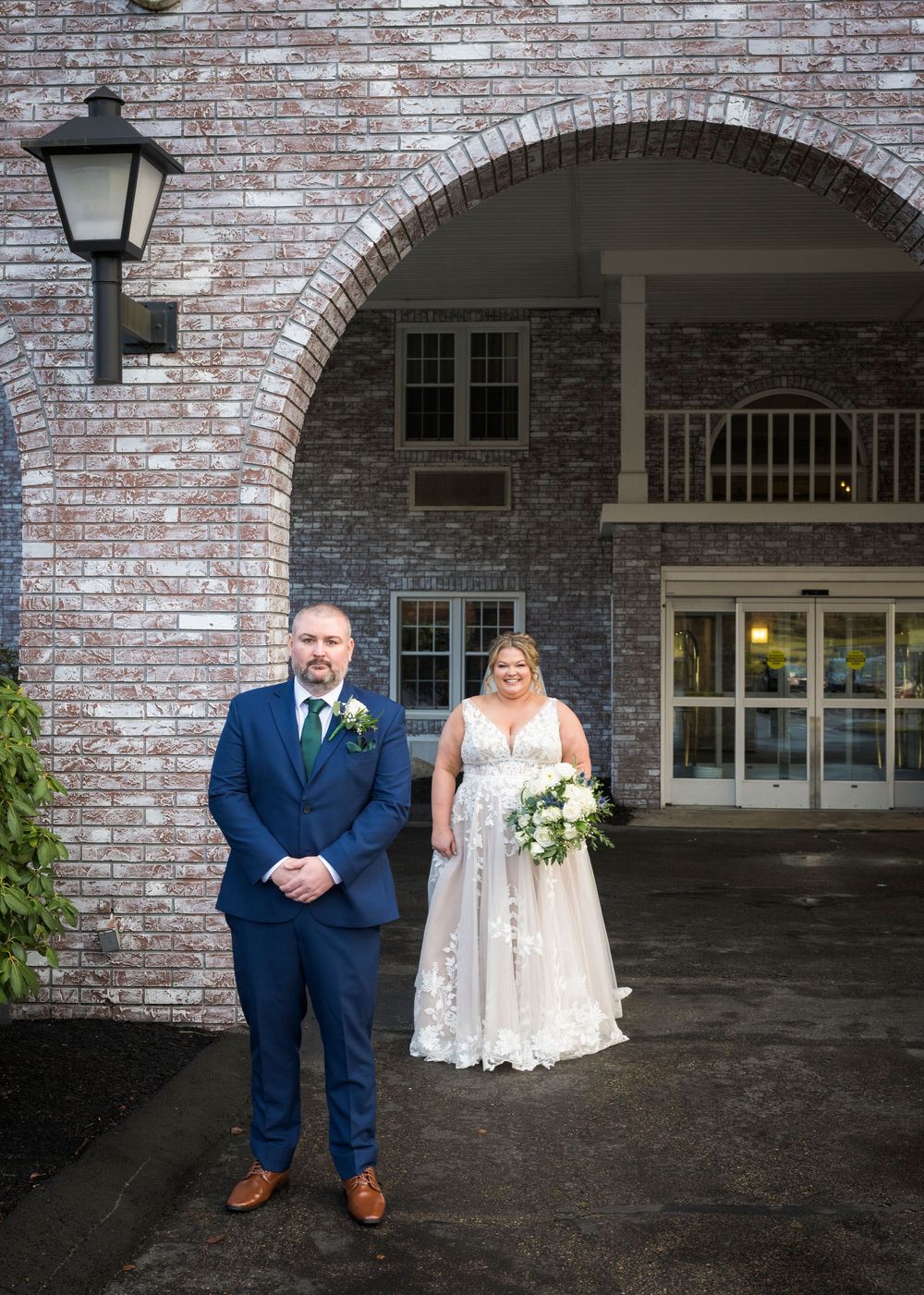 A wedding couple poses under a brick archway with a lantern, featuring a navy suit and white lace gown.