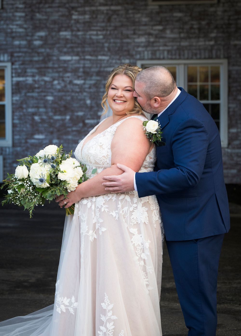 A romantic wedding moment captured against a brick wall with a beautiful bouquet of white roses.