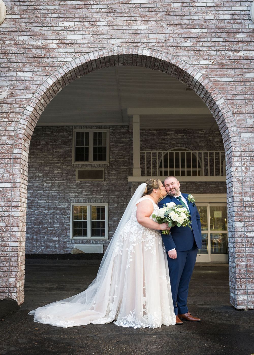A wedding couple shares an intimate moment under a grand brick archway entrance.