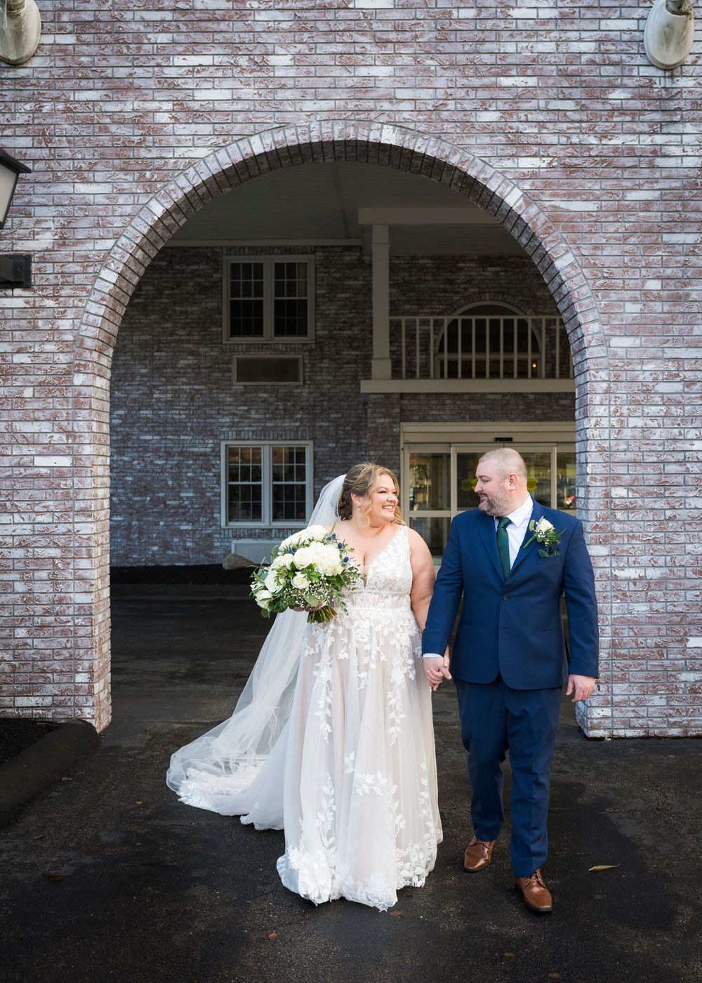 A couple in wedding attire stands beneath a brick archway entrance with white floral bouquet.