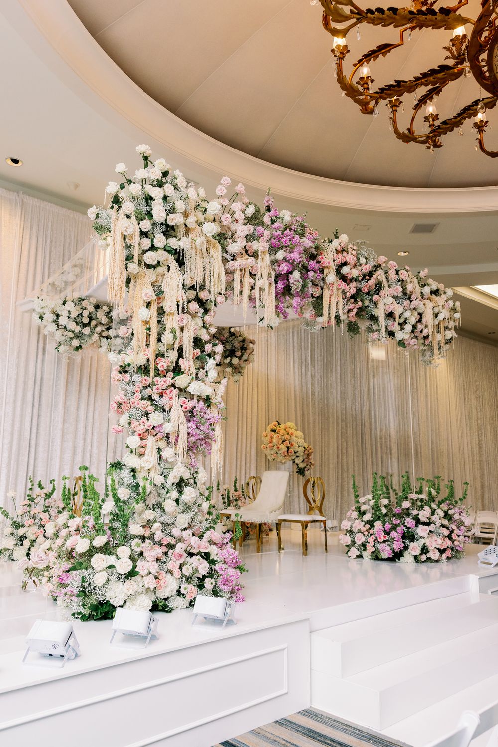 Elegant white and floral wedding stage backdrop with cascading flowers and crystal curtains in a luxurious banquet hall.