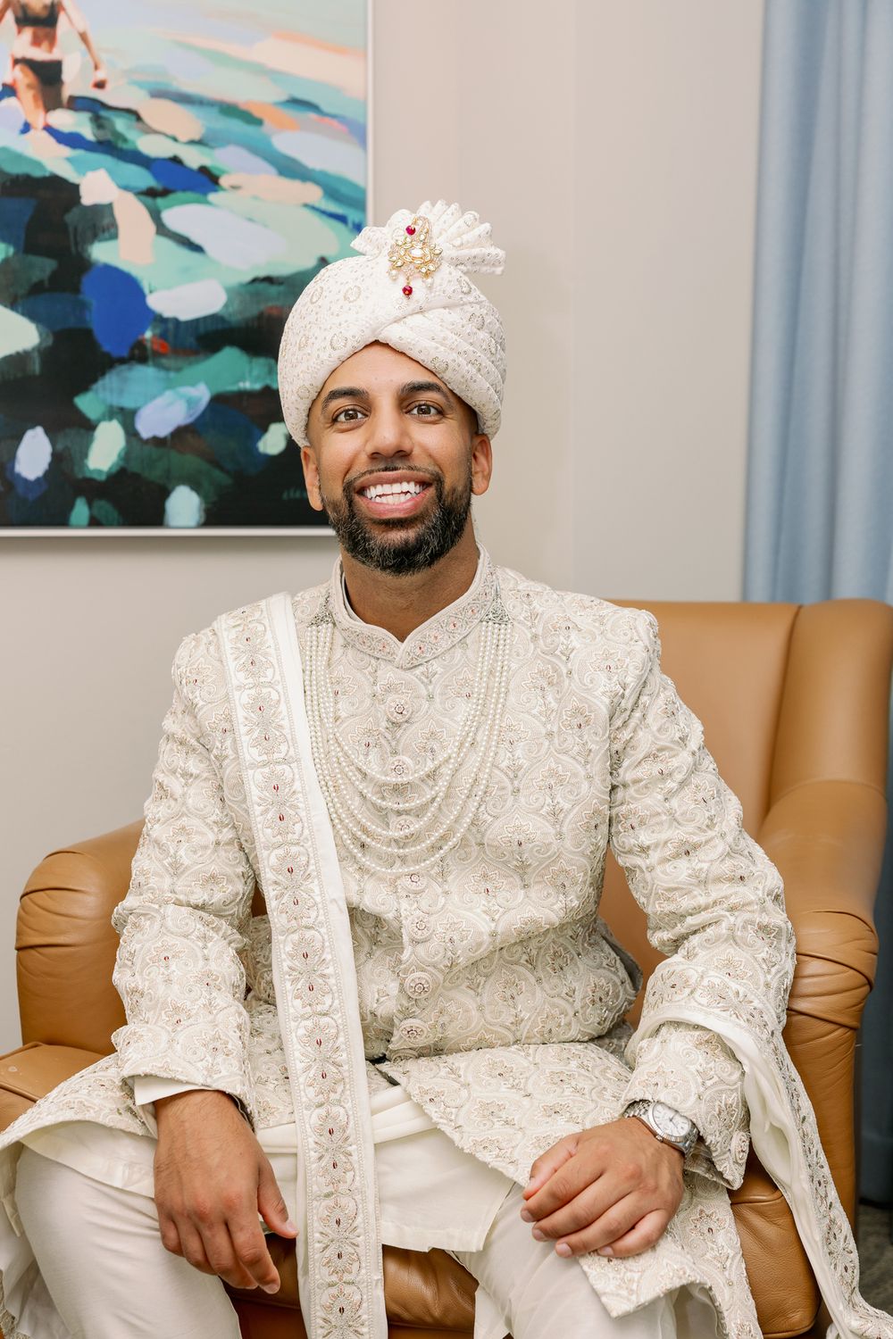 A groom wearing an ornate white sherwani and turban sits on a tan leather couch with decorative artwork in the background.