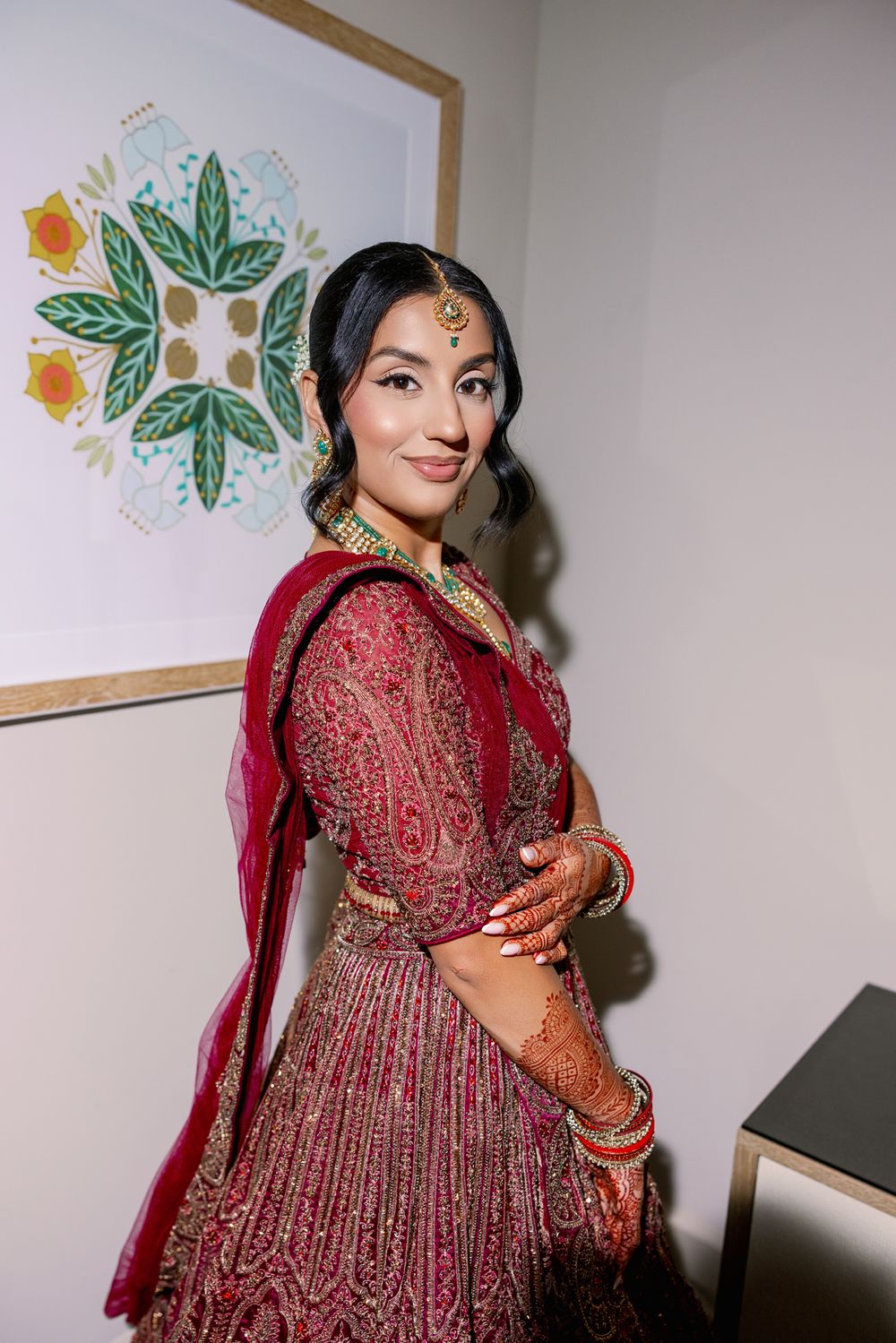 A woman wearing an ornate red and gold lehenga poses elegantly against a wall with floral artwork.