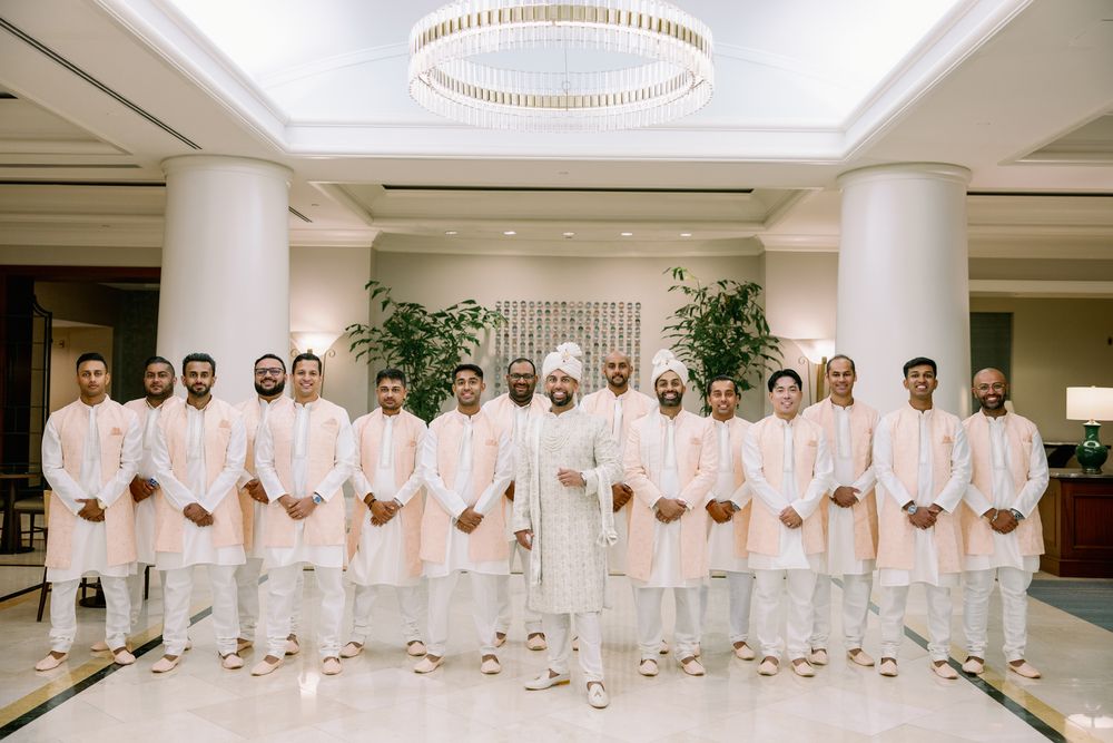 Large group in all-white attire standing in formation for a formal wedding party photo in an indoor venue.