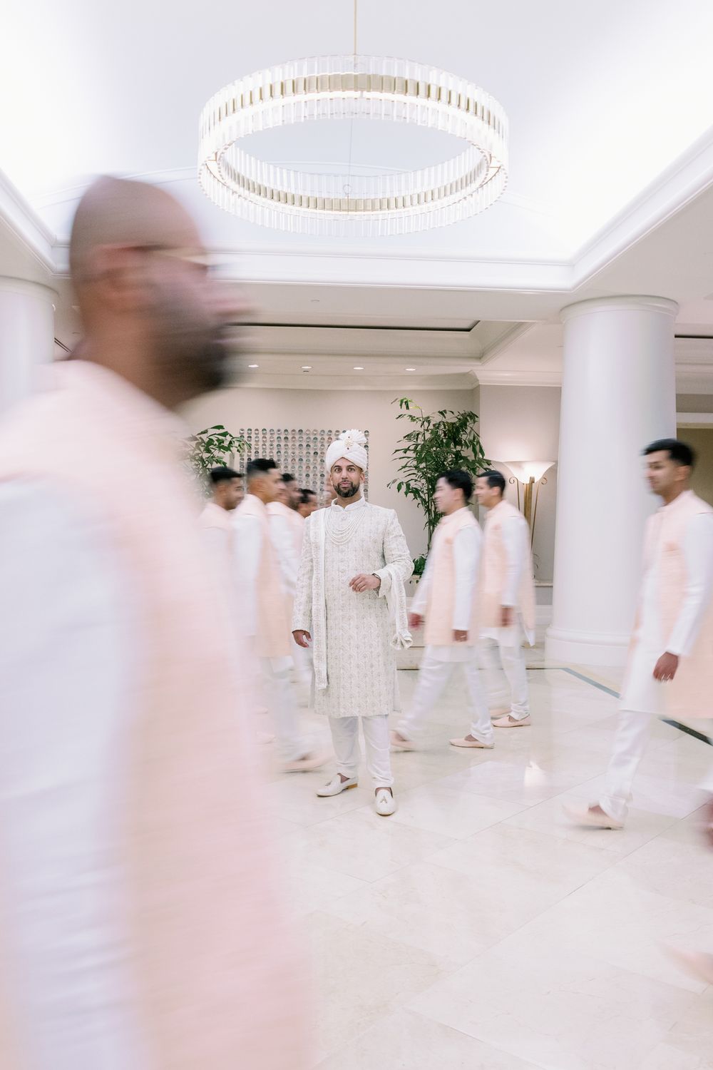 Motion-blurred wedding party in white traditional attire gathered under an elegant circular chandelier.