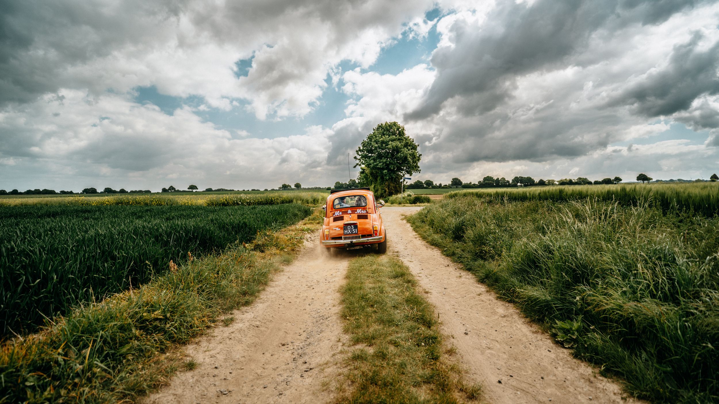 Een bruidspaar vertrekt in een stofwolk over een zandweg met hun klassieke Fiat 500 naar de ceremonie