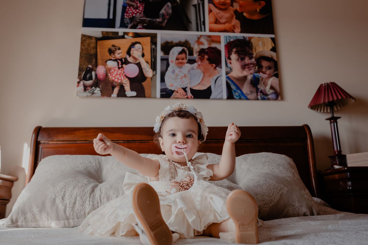 A happy baby in a white dress sitting on a bed with photo frames displayed on the wall.