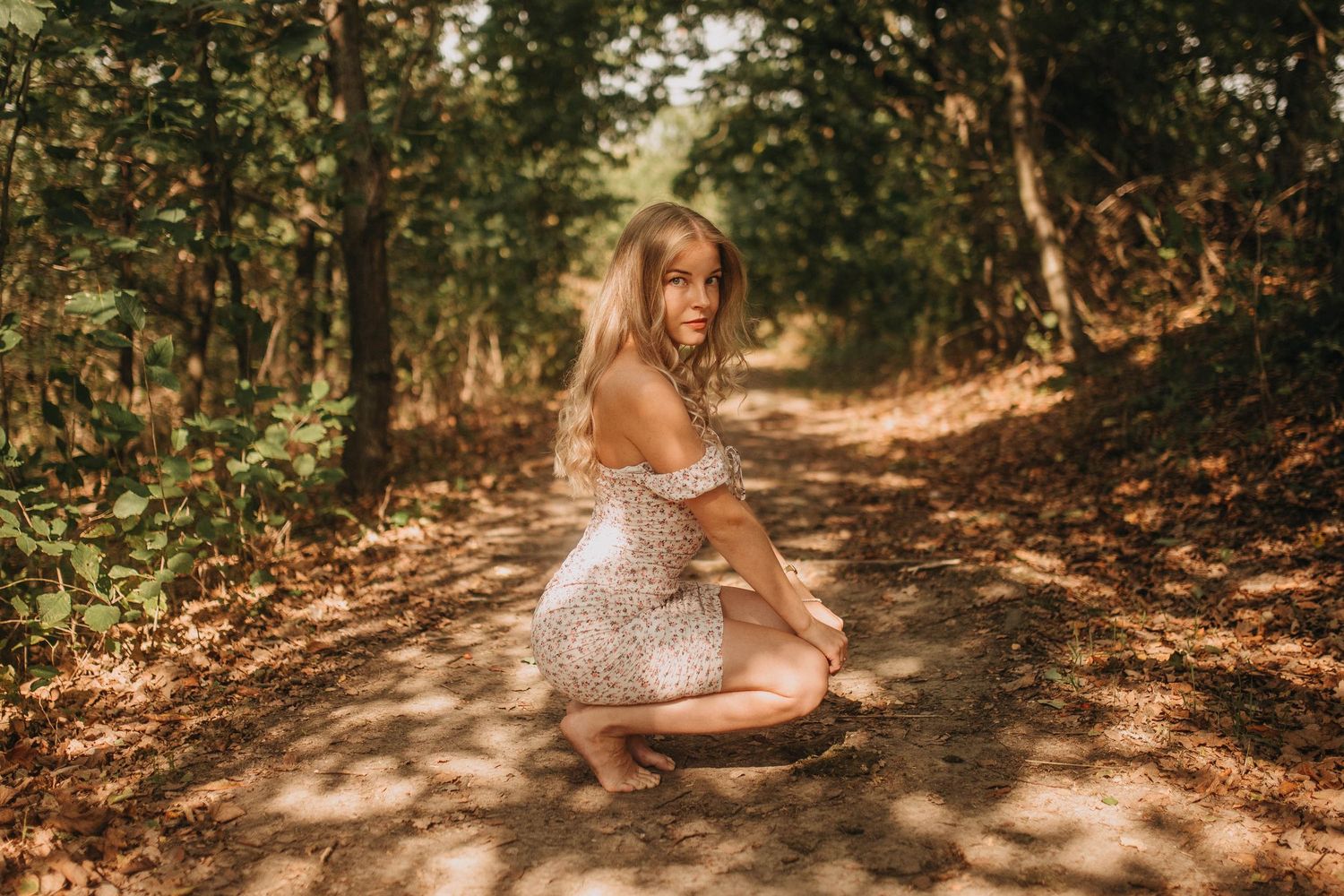 A figure in a sparkly dress poses on a sunlit forest path surrounded by autumn foliage.