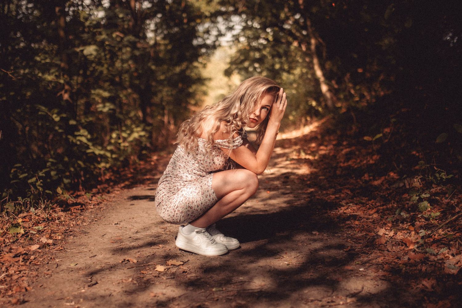 Crouching pose in a sequined dress and white sneakers on a forest path.