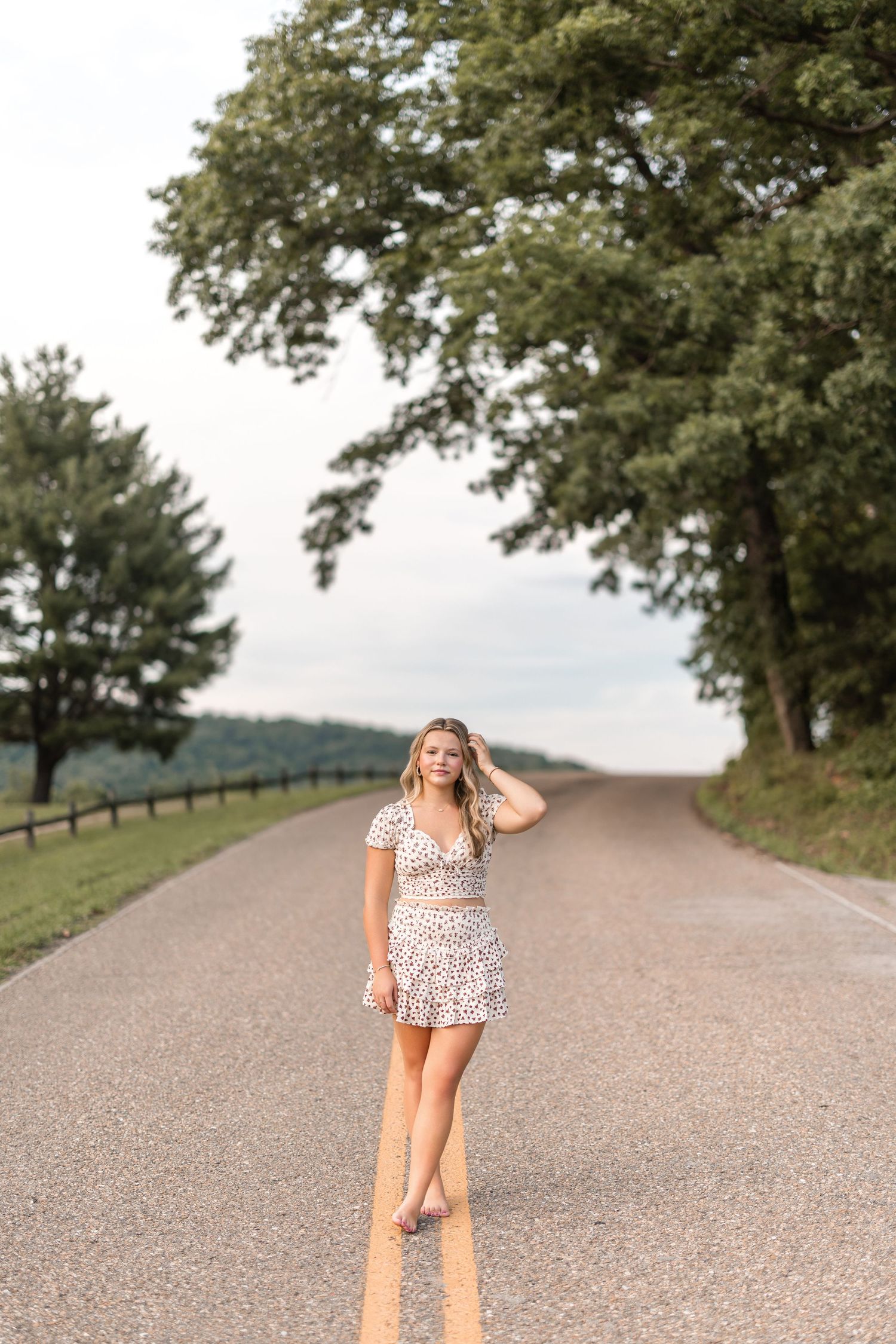 A person in a floral dress poses on a gravel country road surrounded by green trees on a summer day.
