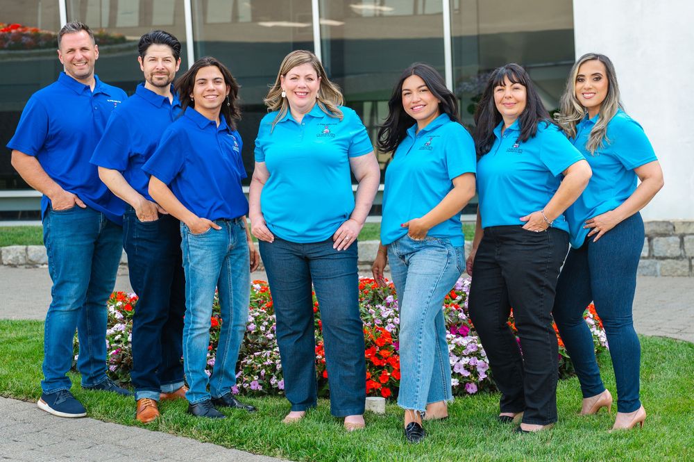 Healthcare workers in blue scrubs and uniforms sit together outdoors for a group photo.