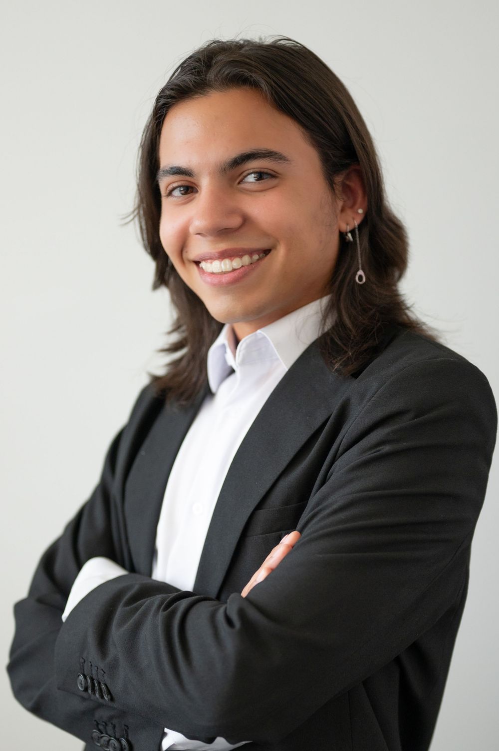 Professional headshot of a person with long dark hair wearing a black blazer and white shirt, smiling with arms crossed.