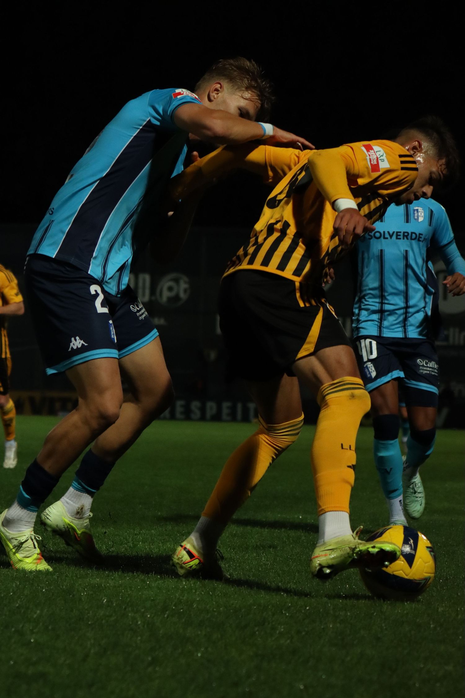 Players in yellow and blue jerseys collide during a physical soccer match under stadium lights.