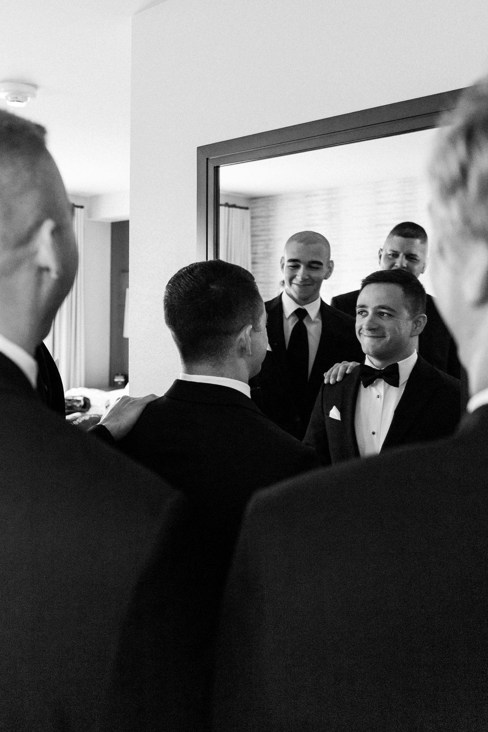 Wedding party members in formal black tuxedos share a candid moment in a bright indoor space.