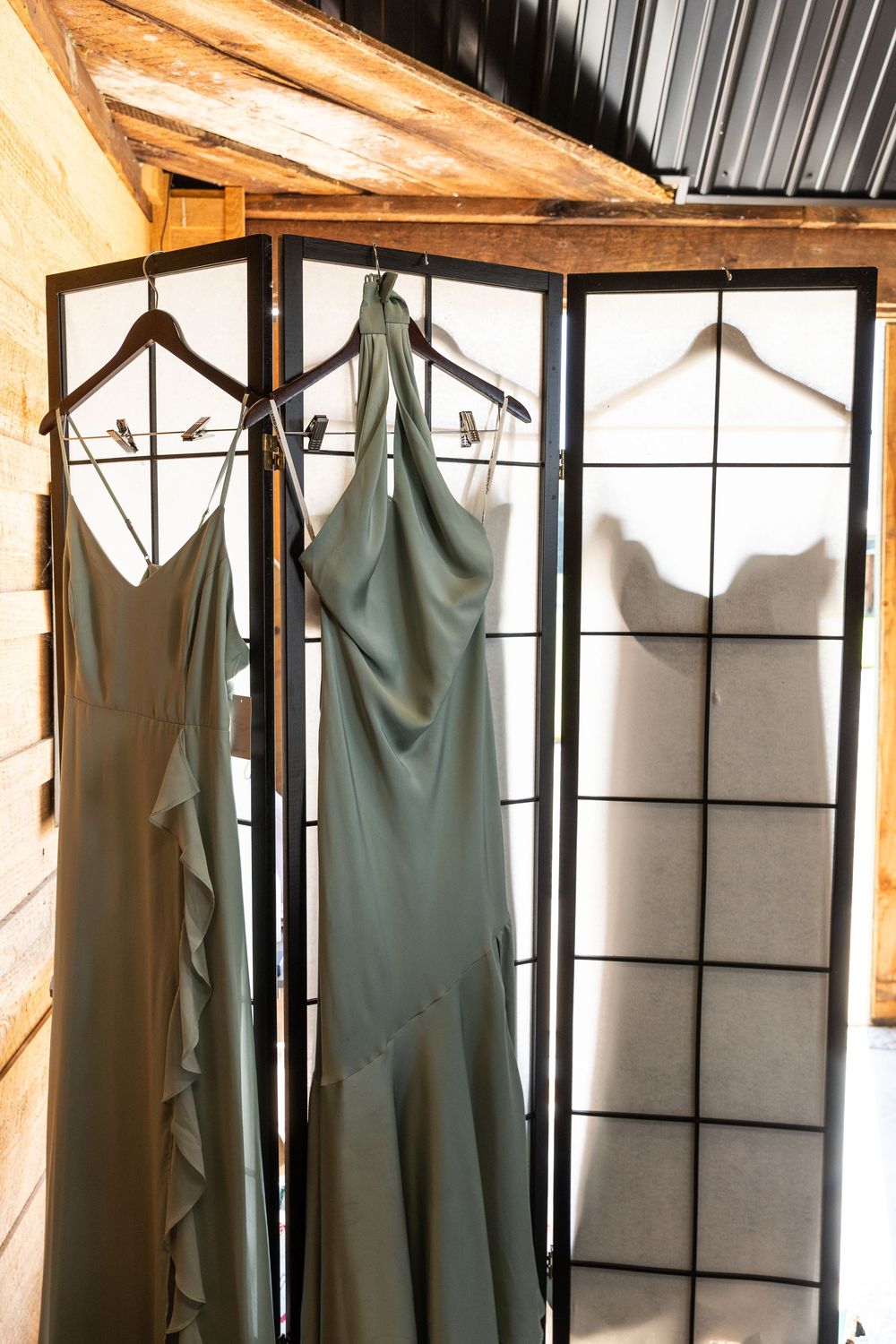 Sage green bridesmaid dresses hanging on wooden hangers in a rustic dressing room.