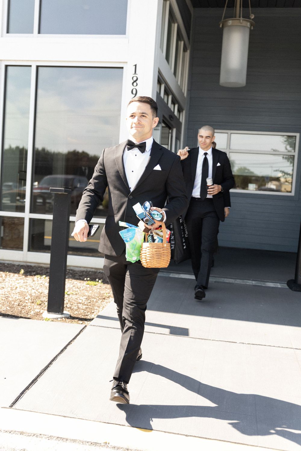 Groomsmen in formal attire carrying gift baskets outside modern building.