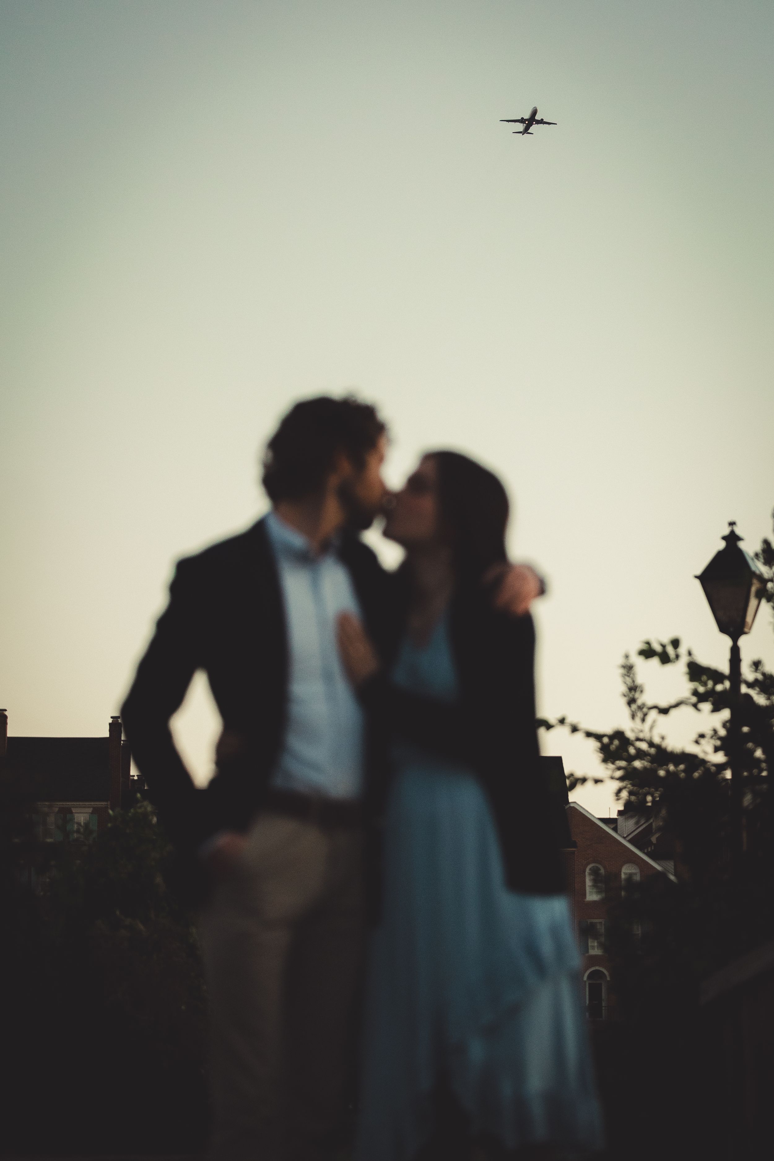A couple in silhouette embraces at dusk as an airplane flies overhead in the background.