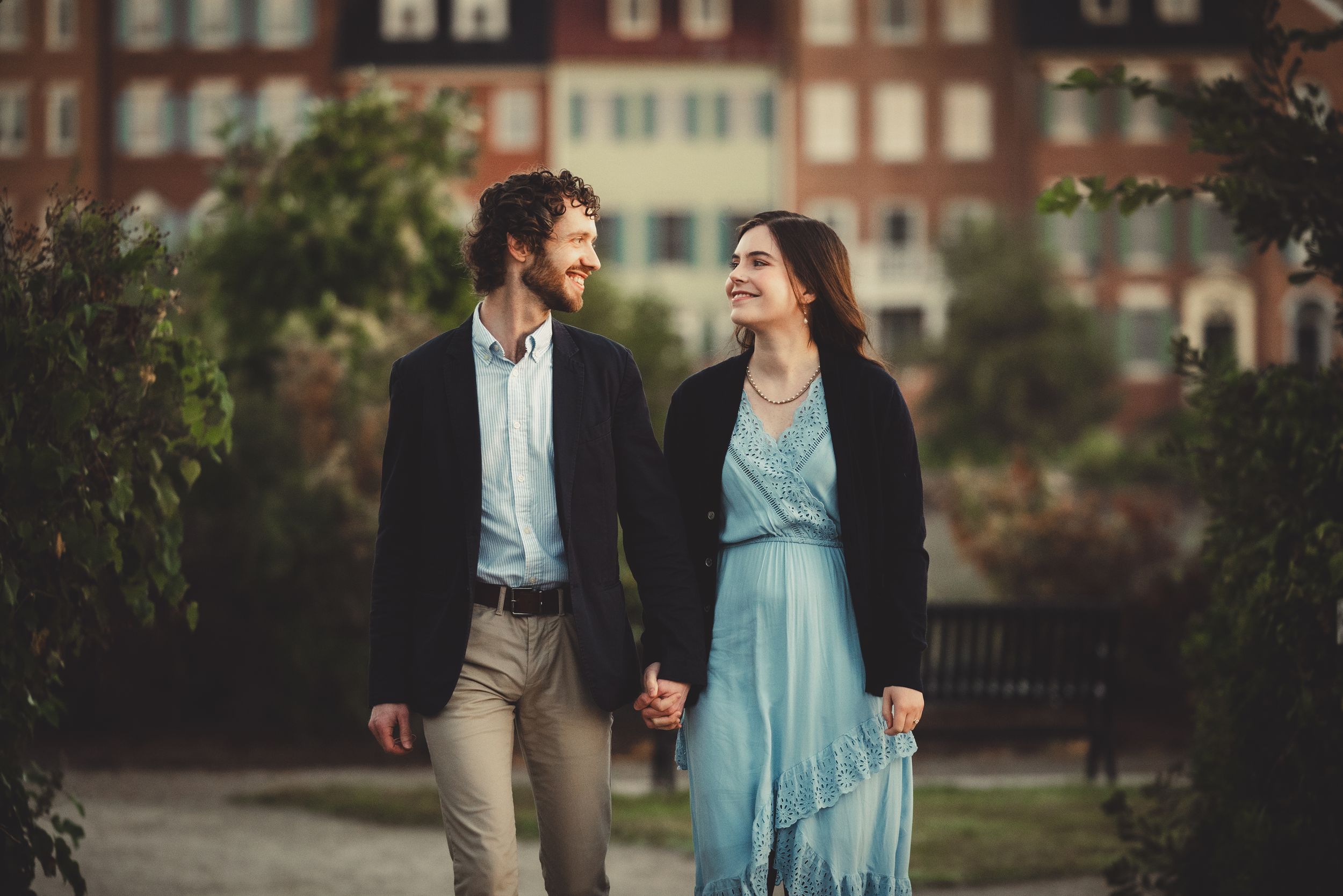 A couple in casual formal attire walk hand in hand along a path with brick buildings in the background at sunset.