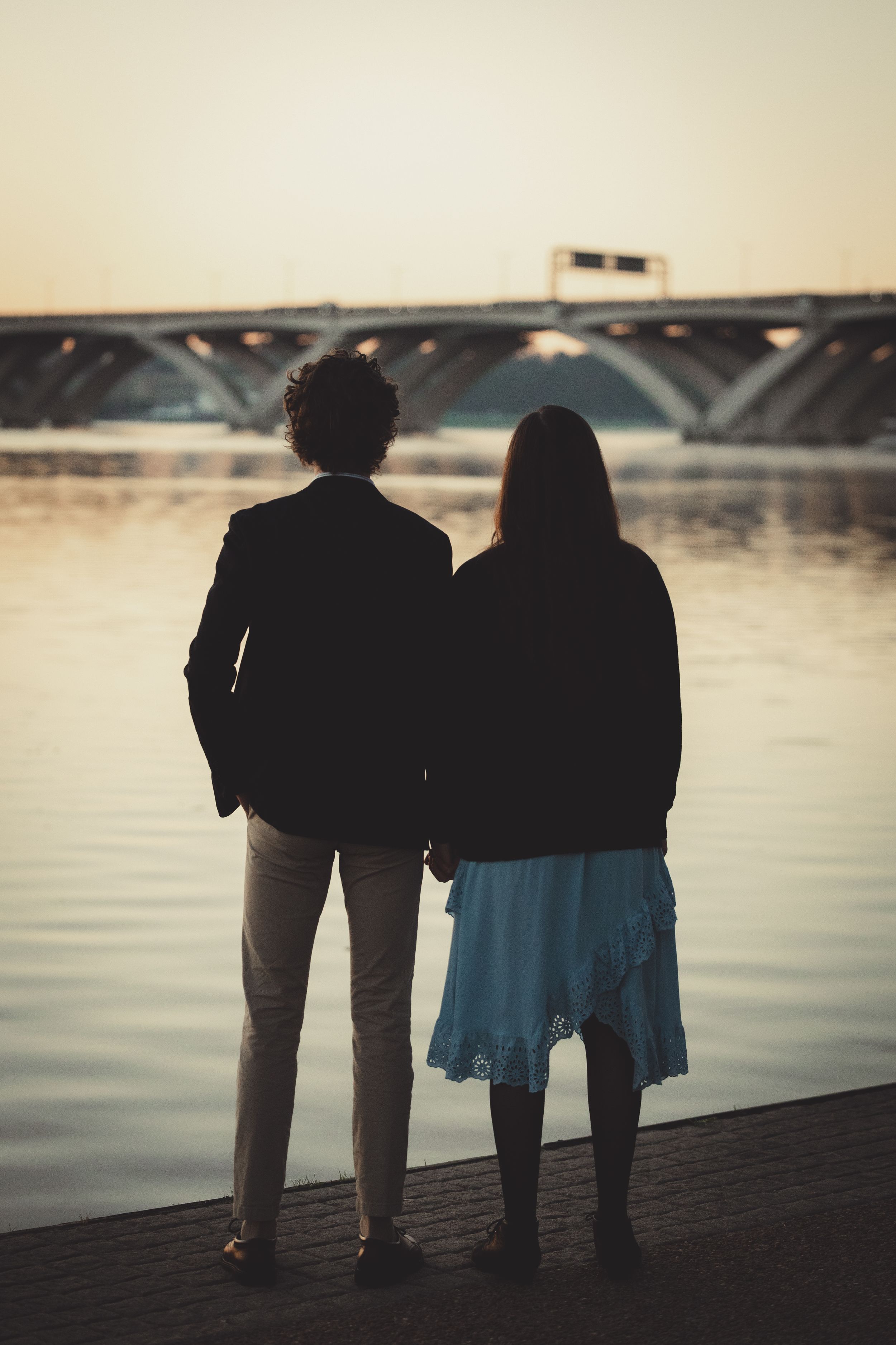 Silhouetted figures stand together on a dock overlooking a serene waterfront at sunset.