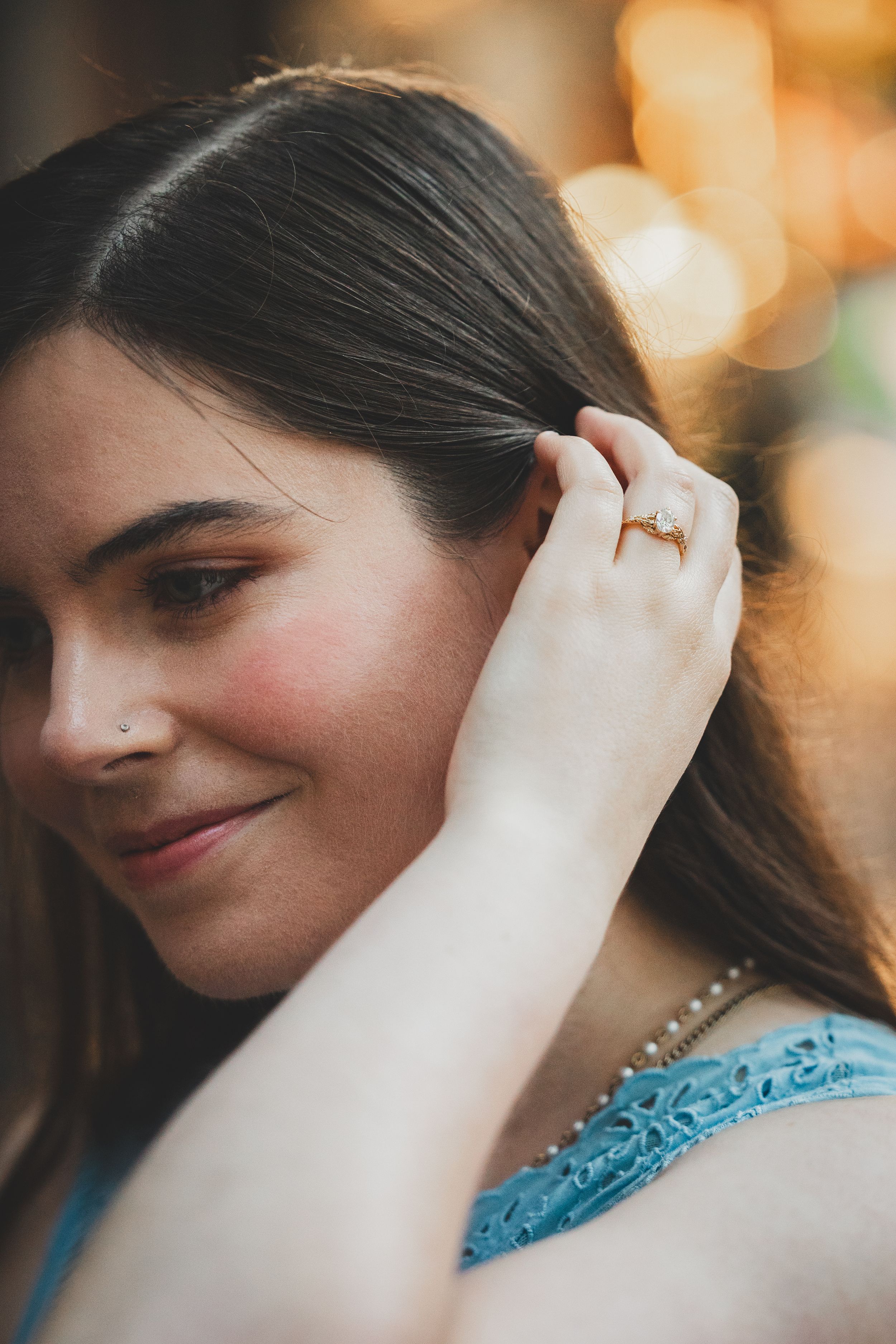 A person wearing a blue lace dress poses with a delicate smile and pearl necklace in warm ambient lighting.