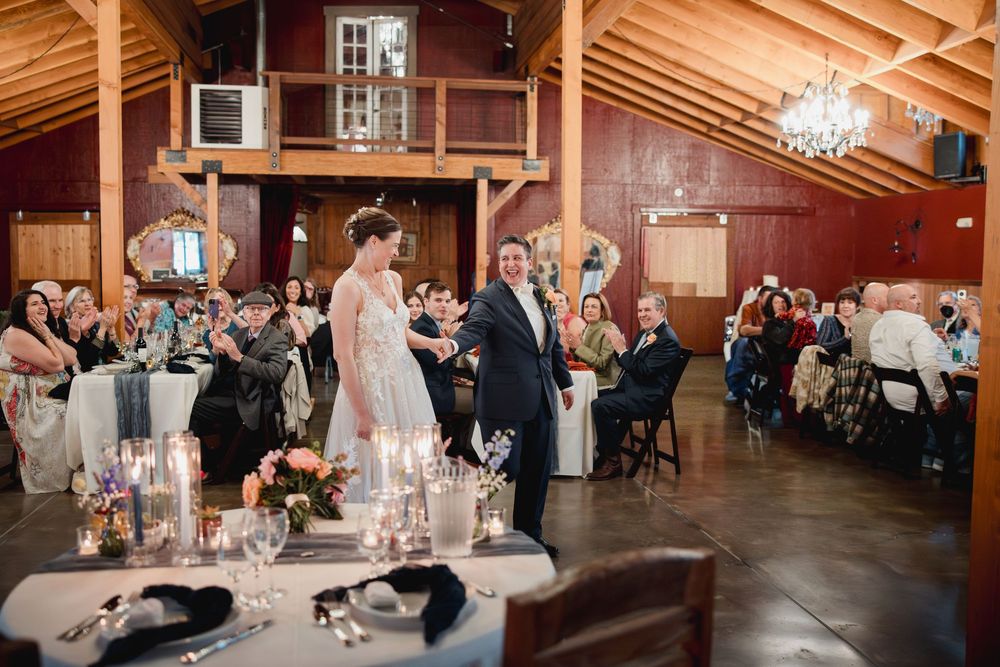 Wedding reception guests dance and celebrate in a rustic barn venue with elegant chandeliers overhead.