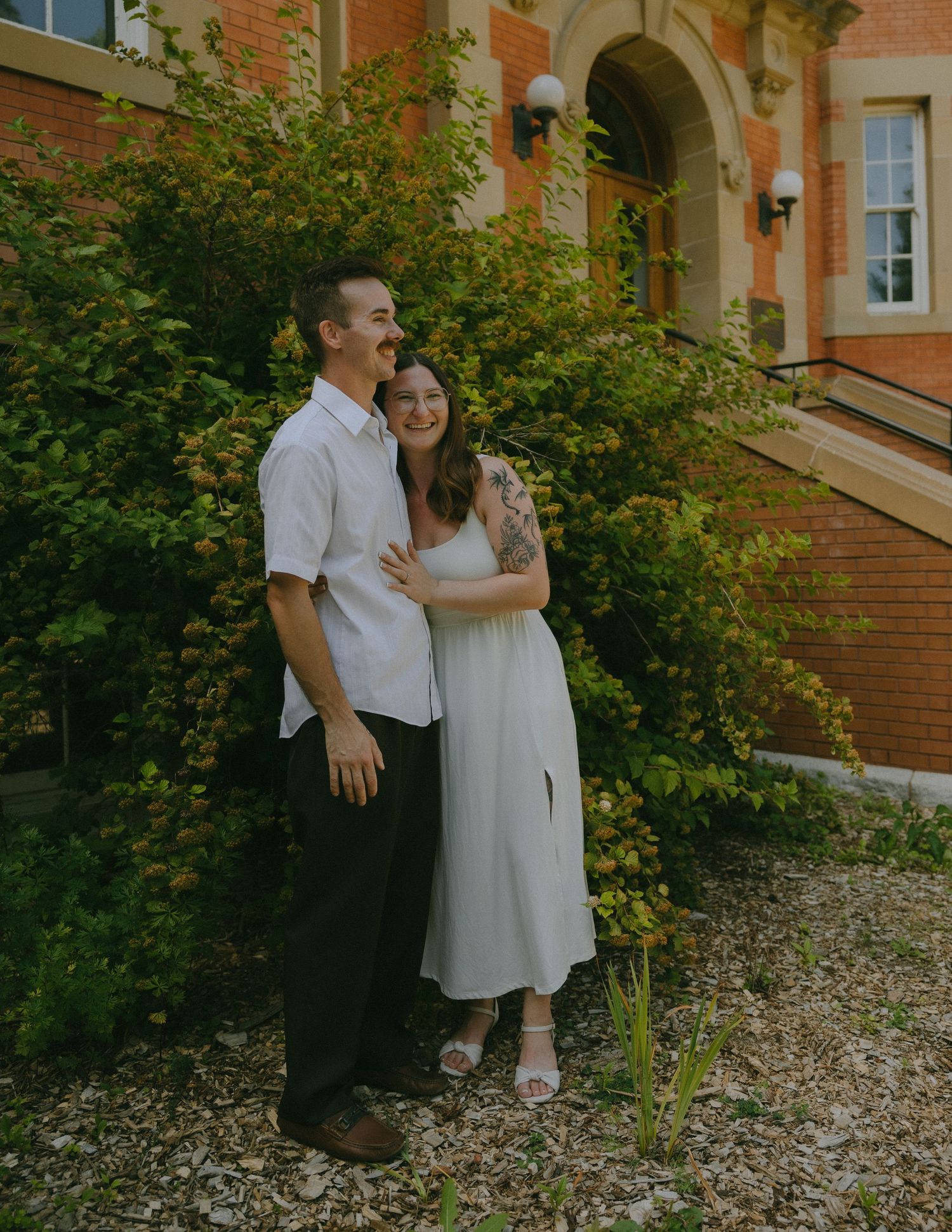 A couple shares romantic moments in front of a brick building surrounded by lush greenery.