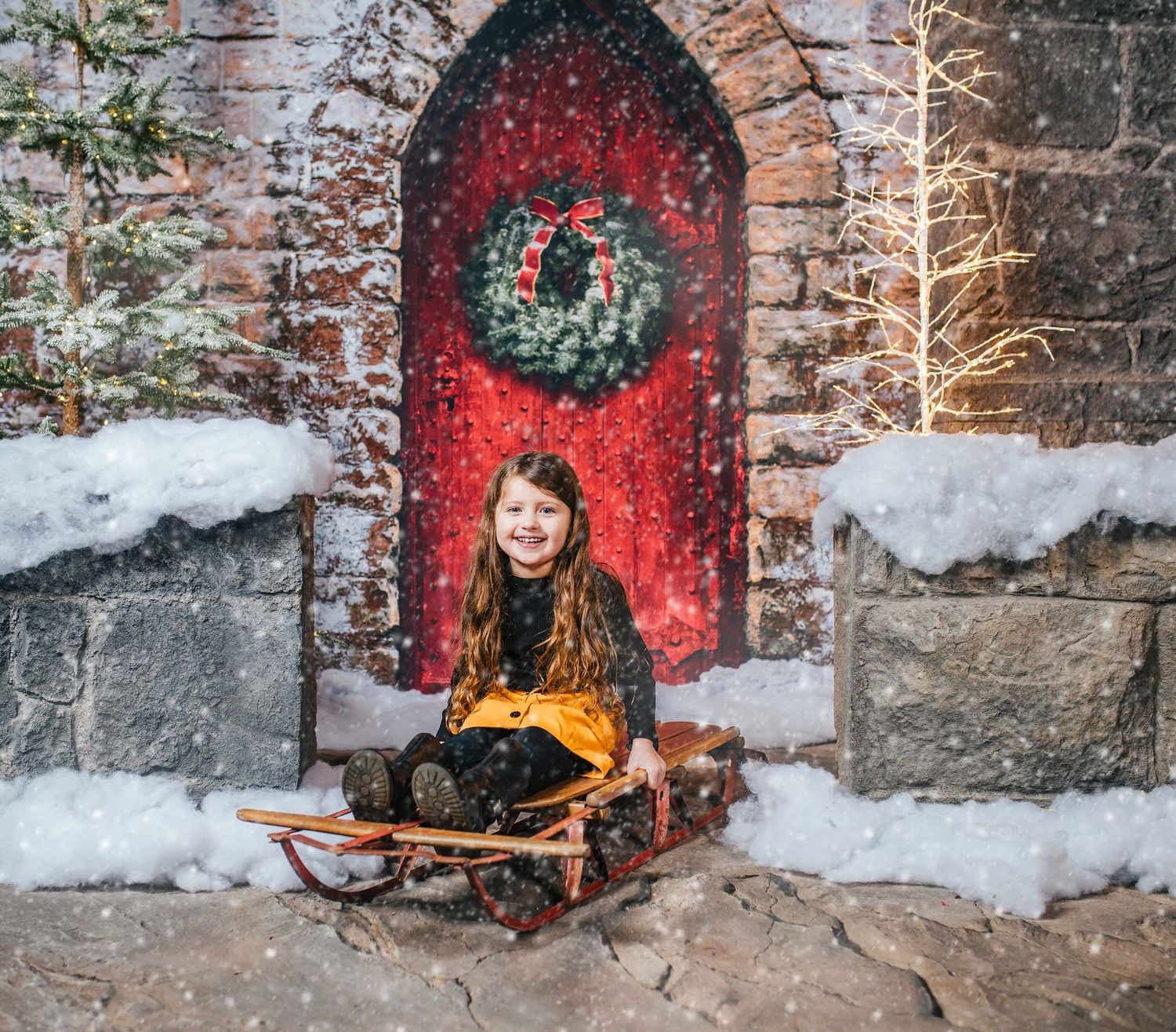 Winter scene with child on sled against red door with wreath and brick archway.
