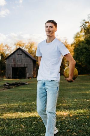 Person in white t-shirt and light jeans holds football in golden sunlight with rustic barn in background.