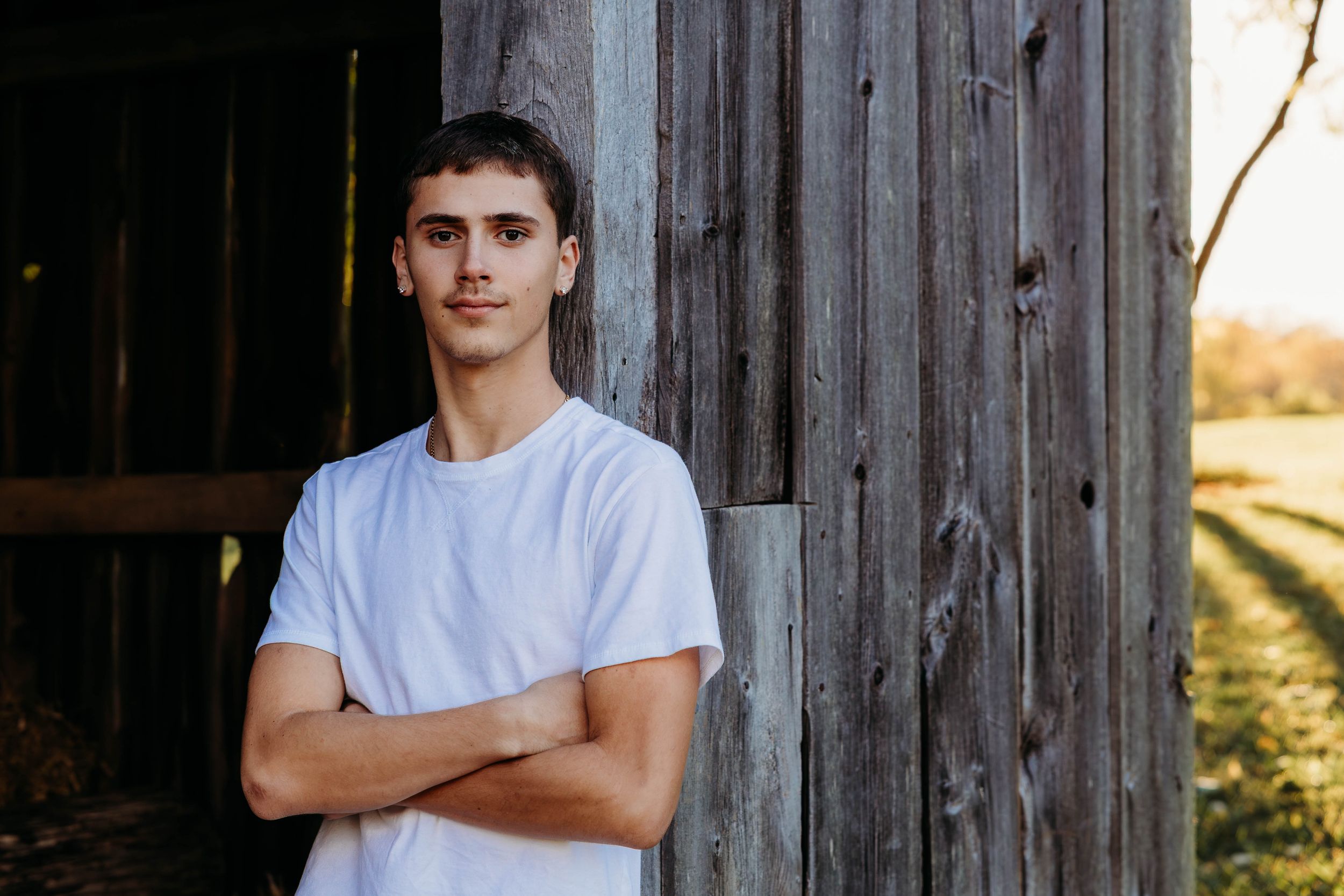 Someone in a white t-shirt stands with arms crossed against a rustic wooden barn wall at sunset.