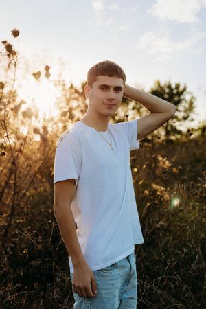 A person in a white t-shirt and jeans poses in a golden-lit field at sunset with tall grass and wildflowers.