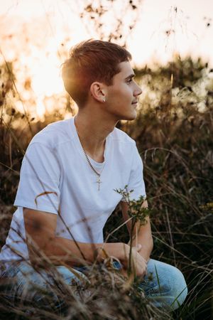 A person in a white t-shirt sits thoughtfully in tall grass during golden hour sunset.