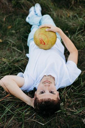 A young child in a white outfit lies in the grass holding a baseball while looking up at the sky.